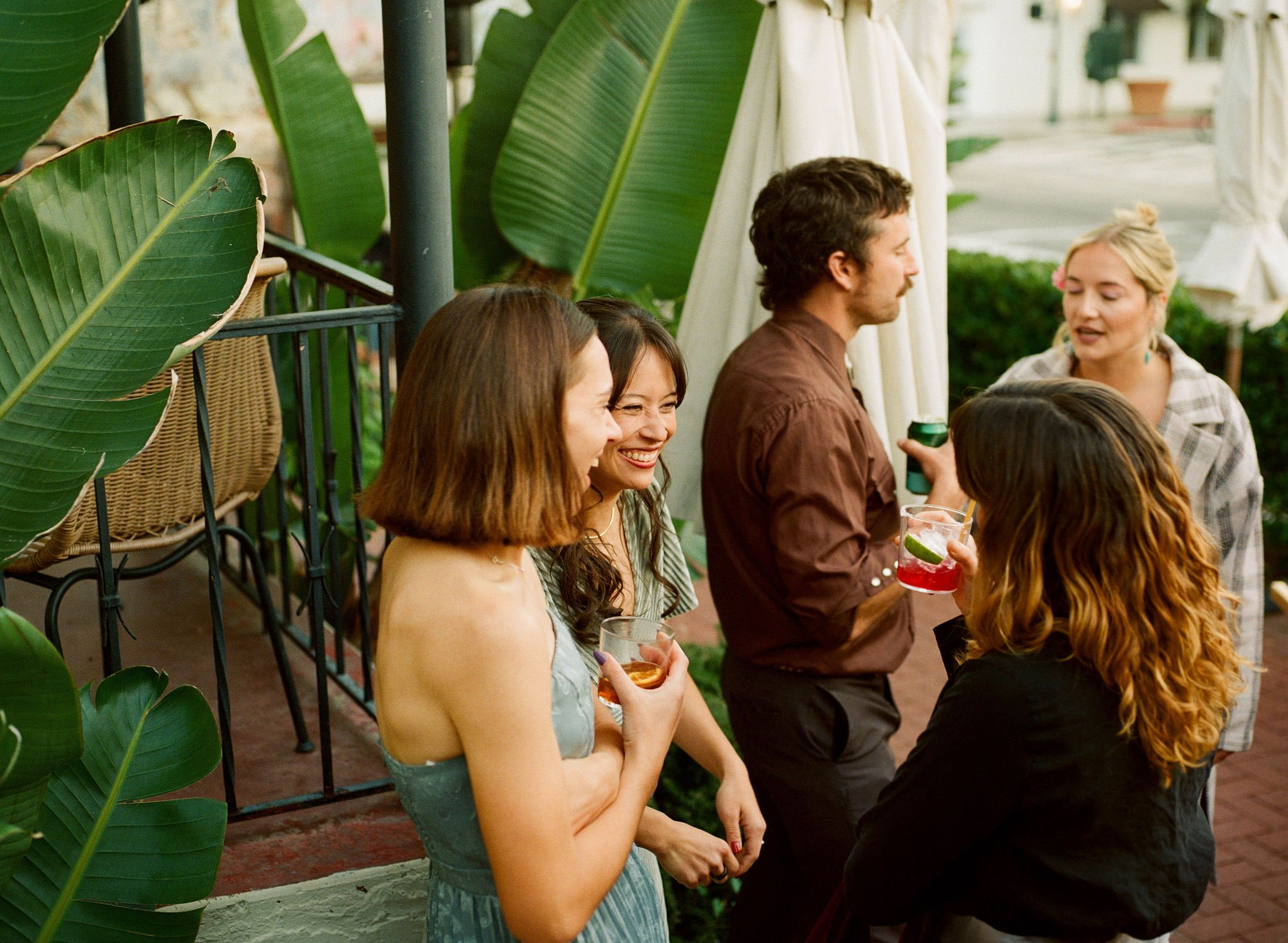 Group of people socializing at an outdoor gathering, some holding drinks, with green plants and patio umbrellas in the background.