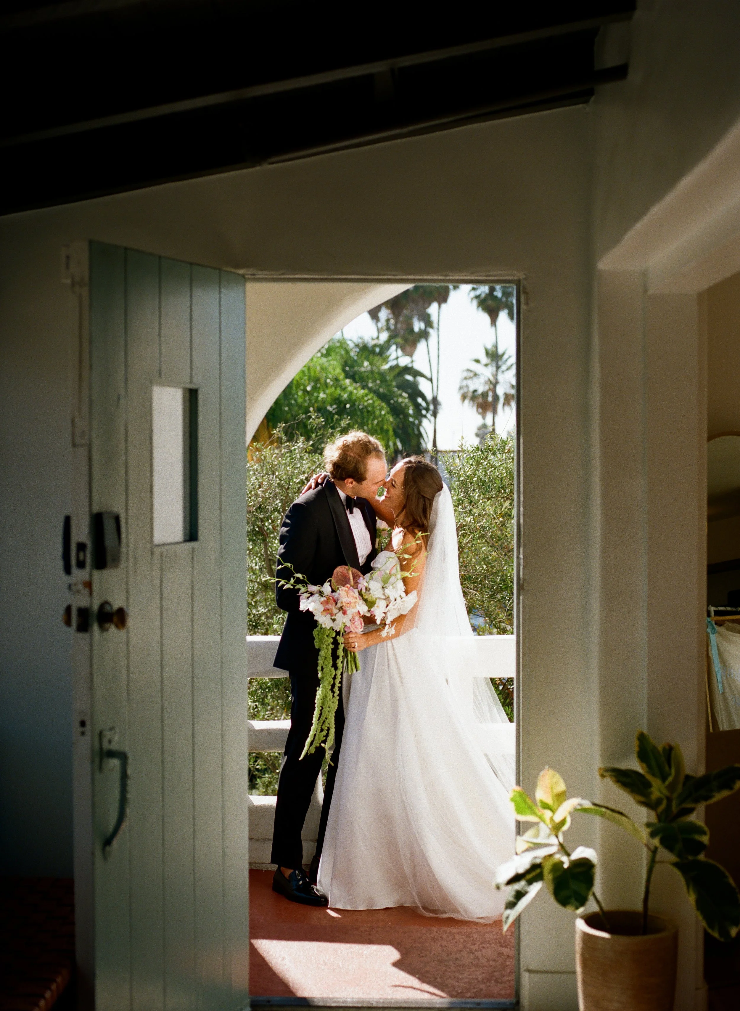A bride and groom share a kiss on a sunlit porch, framed by an open door, with palm trees and greenery in the background.
