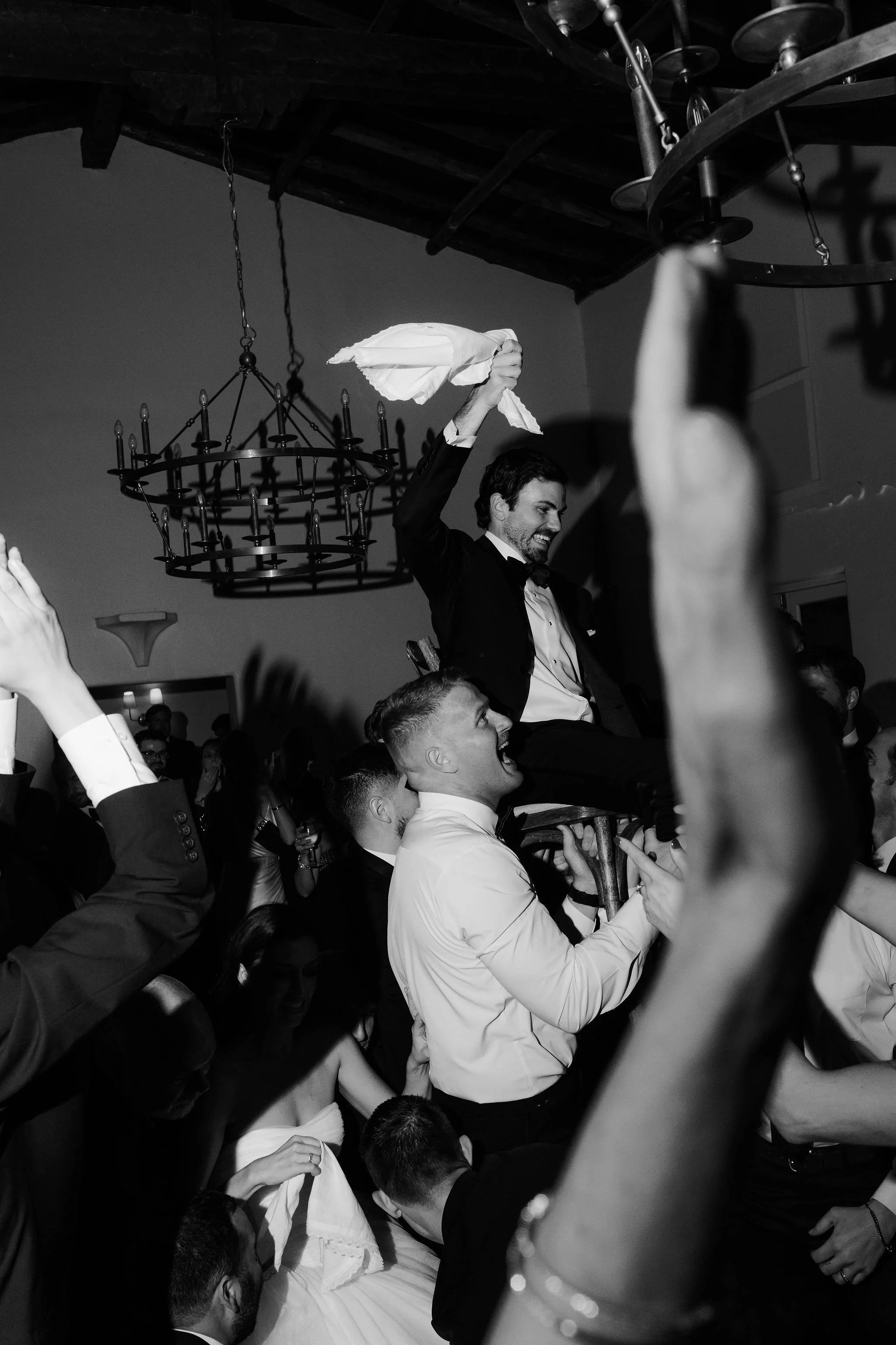 People at a wedding celebration, some are dancing and raising their hands, a man in a tuxedo is holding a cloth and smiling, a chandelier is hanging from the ceiling.