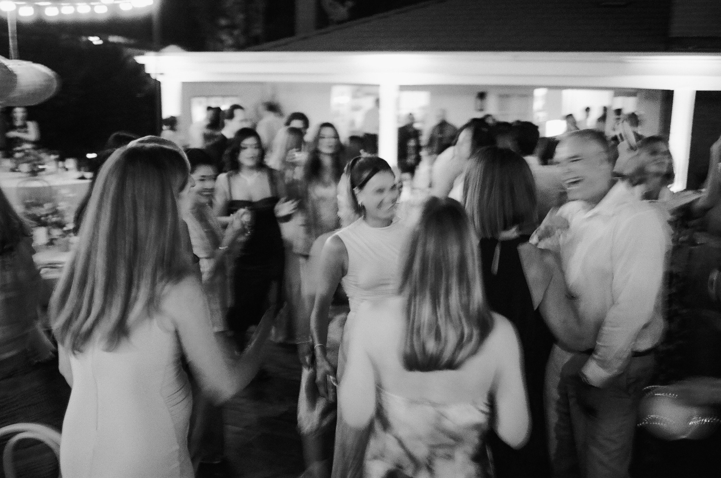 A group of women dressed in white are gathered together in a social setting at night, engaging in conversation and smiling.