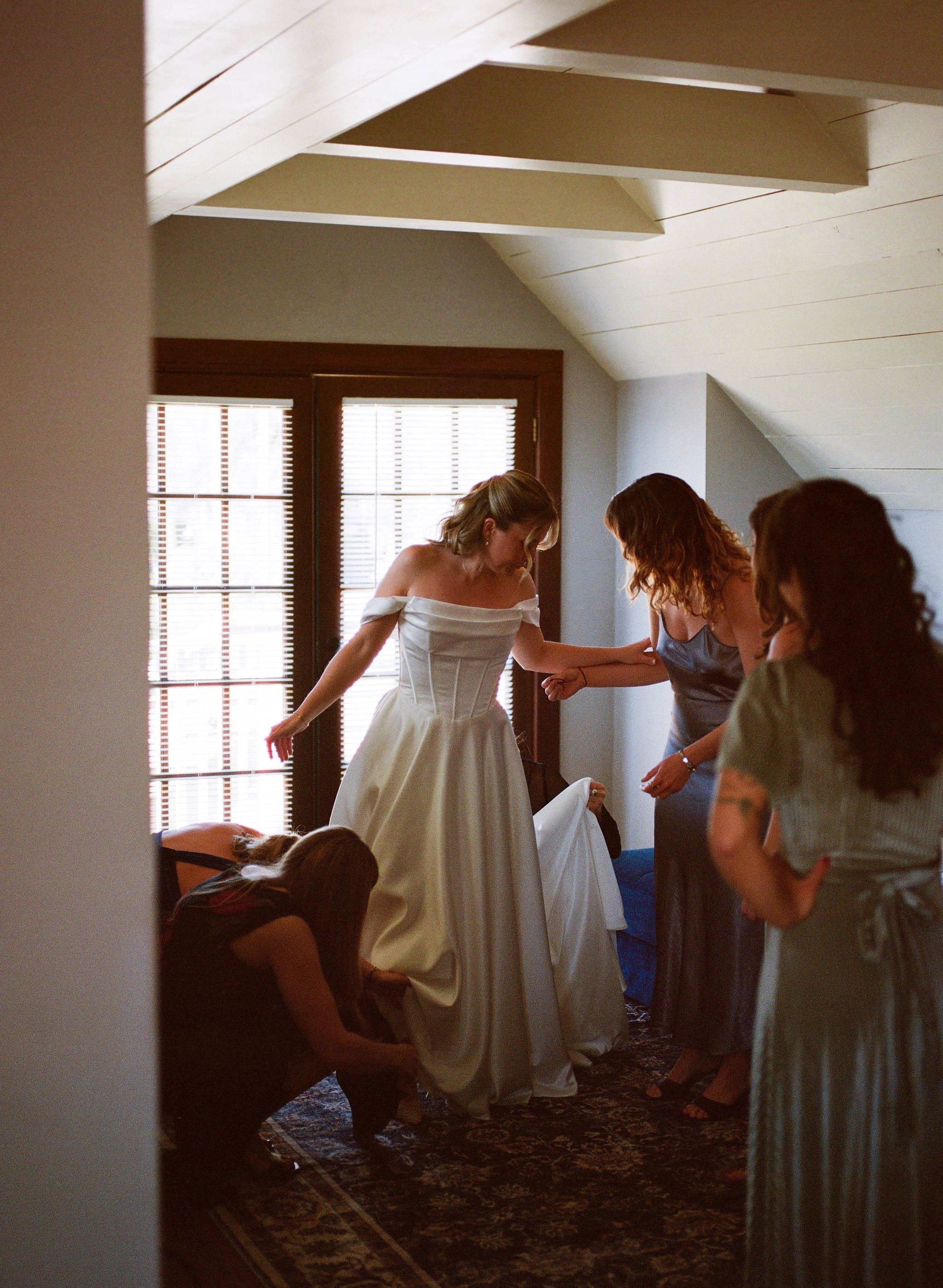 Women preparing a wedding dress in a room with natural light through window blinds, with others helping and one person crouching on the floor.