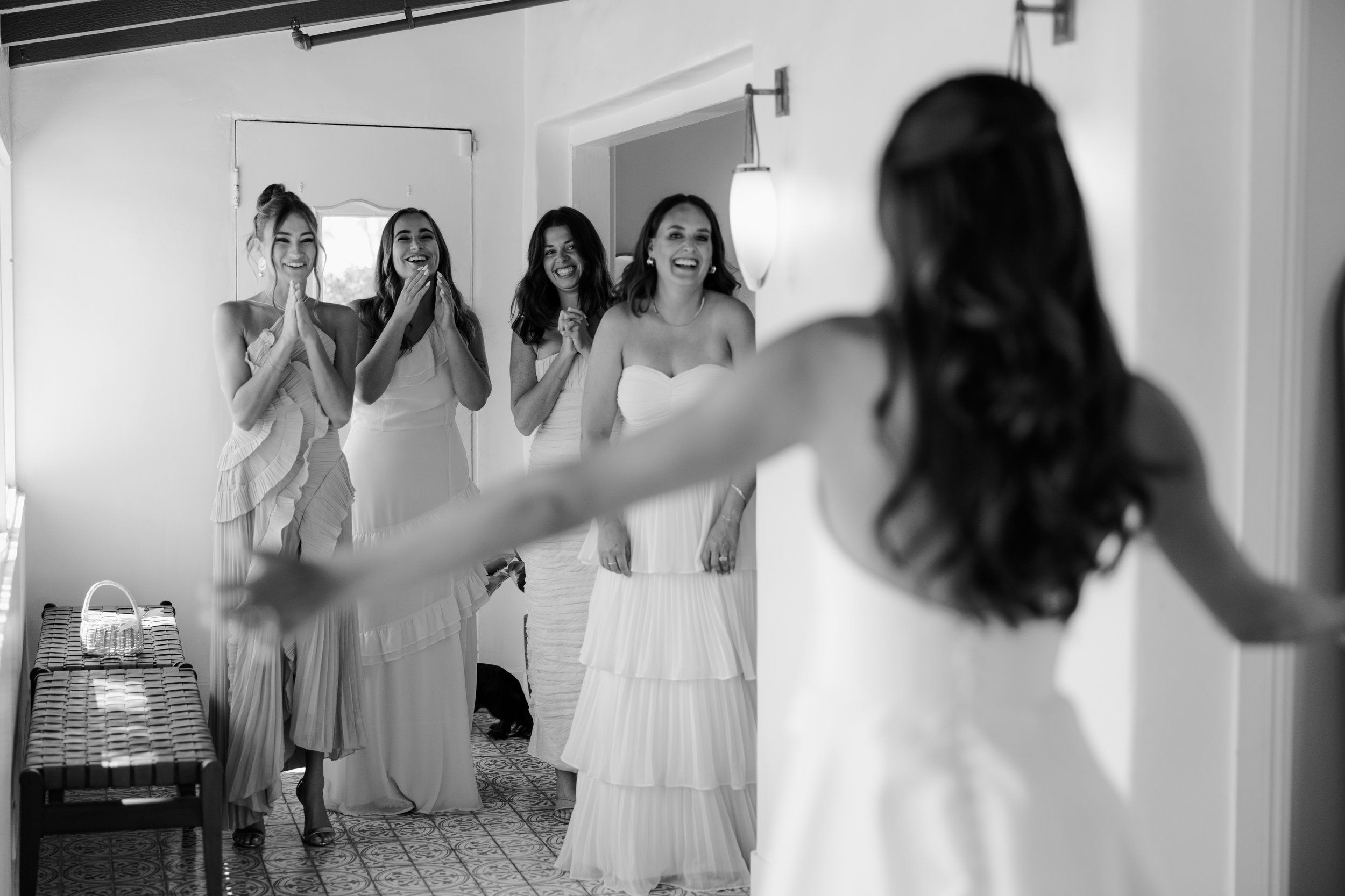 Multiple women dressed in elegant white dresses smiling and laughing in a room, with one woman in the foreground seen from behind, possibly during a bridal preparation or celebration.