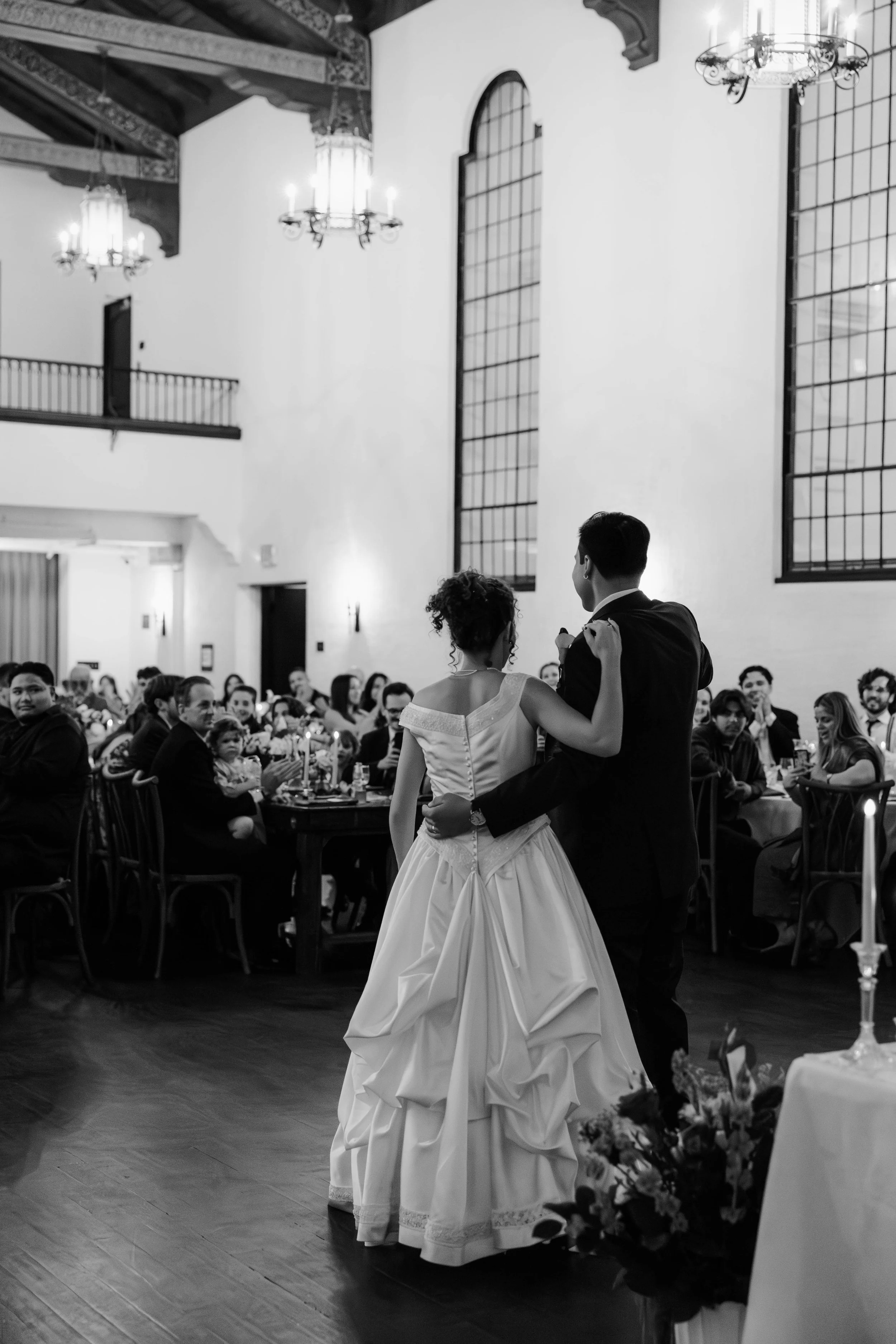 A couple is dancing in front of a seated audience at a wedding reception in an elegant hall with high ceilings, large windows, and chandeliers.