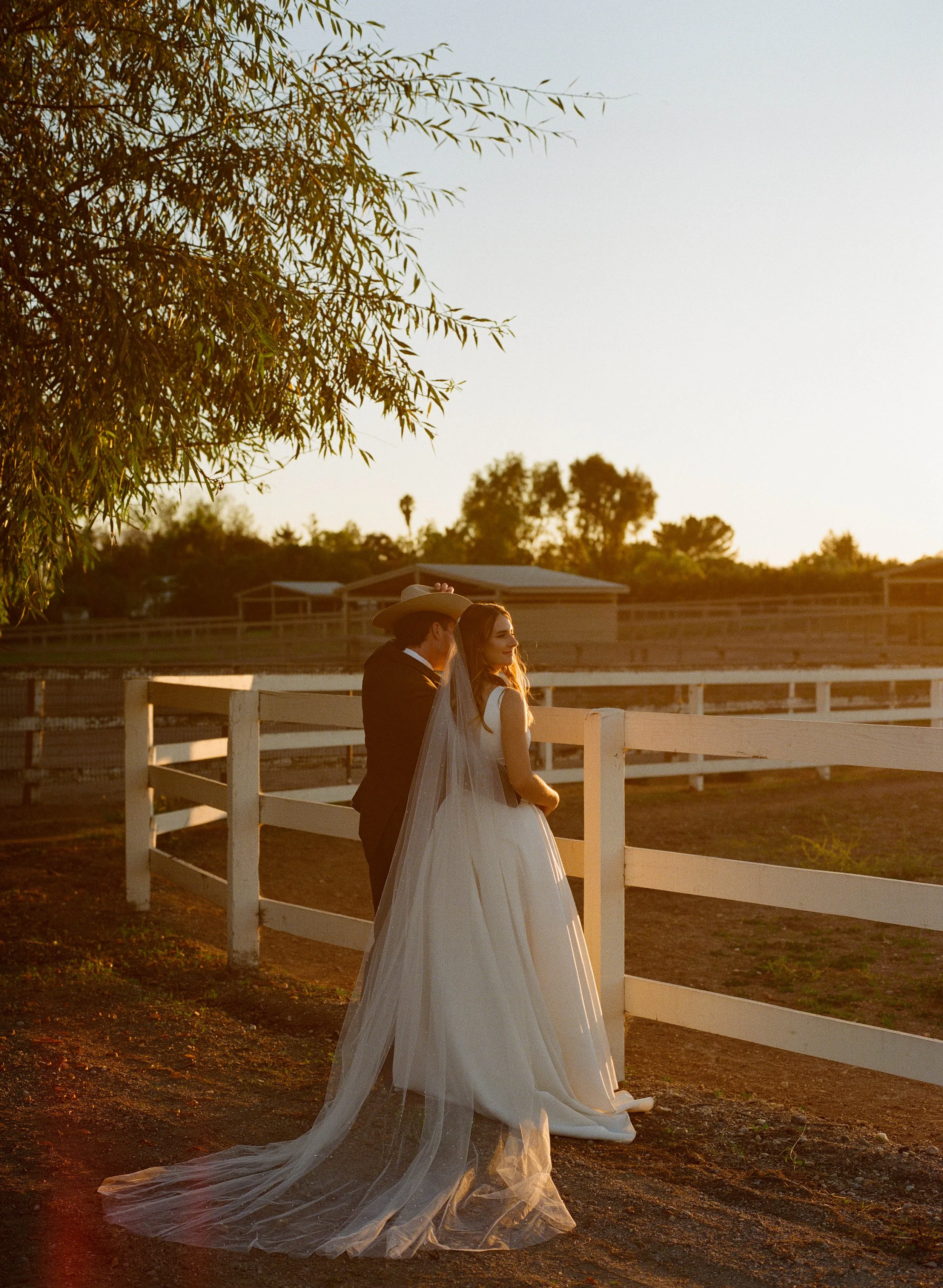 A bride and groom standing outdoors beside a white fence during sunset. The bride is wearing a white wedding gown and veil, and the groom is in a dark suit. They are looking at each other.