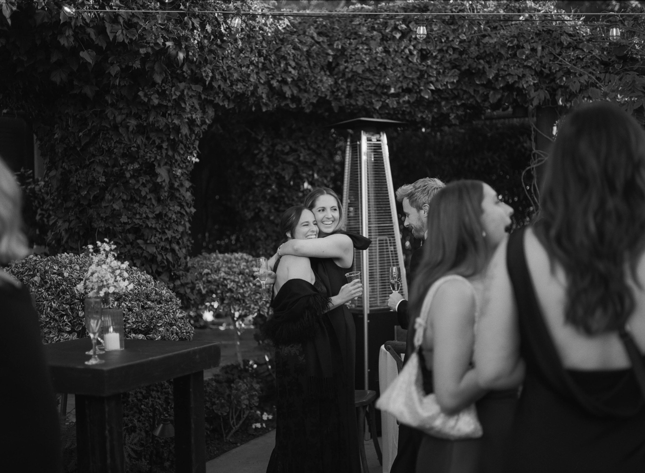People are enjoying a social gathering outdoors in a garden with lush foliage and string lights. They are smiling, hugging, and holding drinks, indicating a festive or celebratory atmosphere.