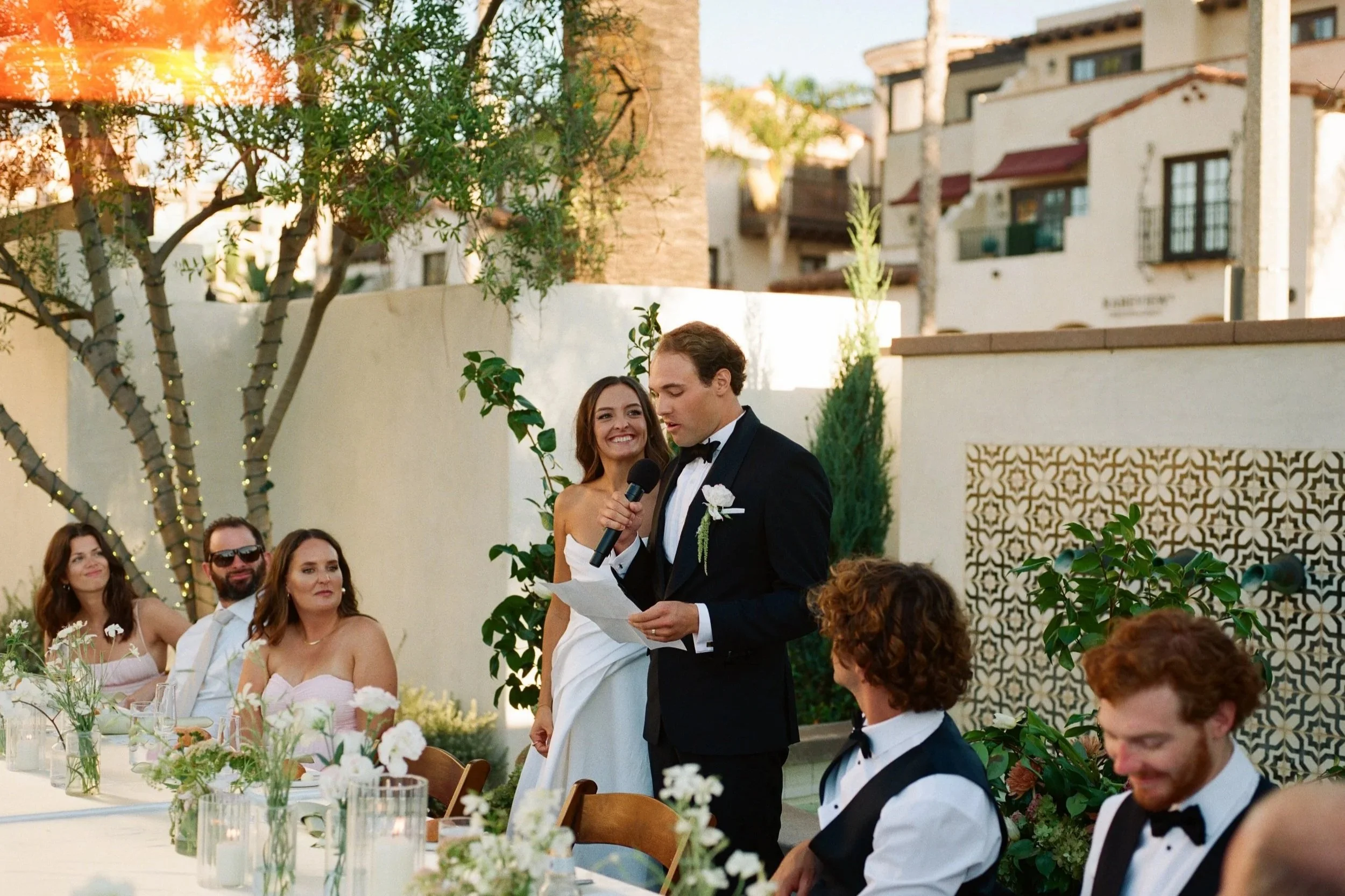 A wedding ceremony outdoors with a groom reading vows, surrounded by guests. The bride and wedding party are present, with the setting featuring trees, string lights, and a modern building in the background.