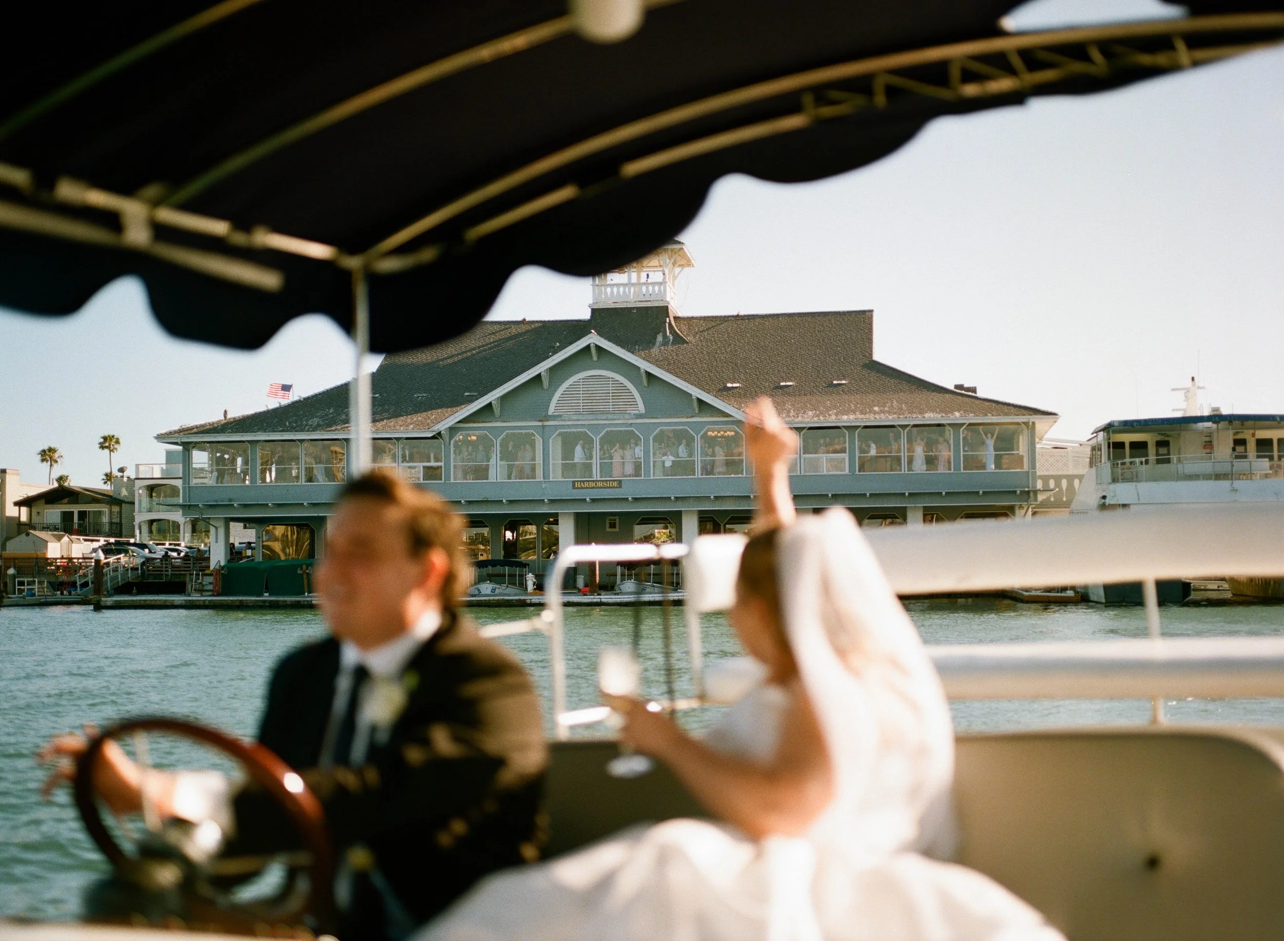 A view of a building labeled 'Harbourside' with a large green canopy overhead, situated near water with boats docked nearby, seen from a boat with a black canopy and a person in formal attire in the foreground.
