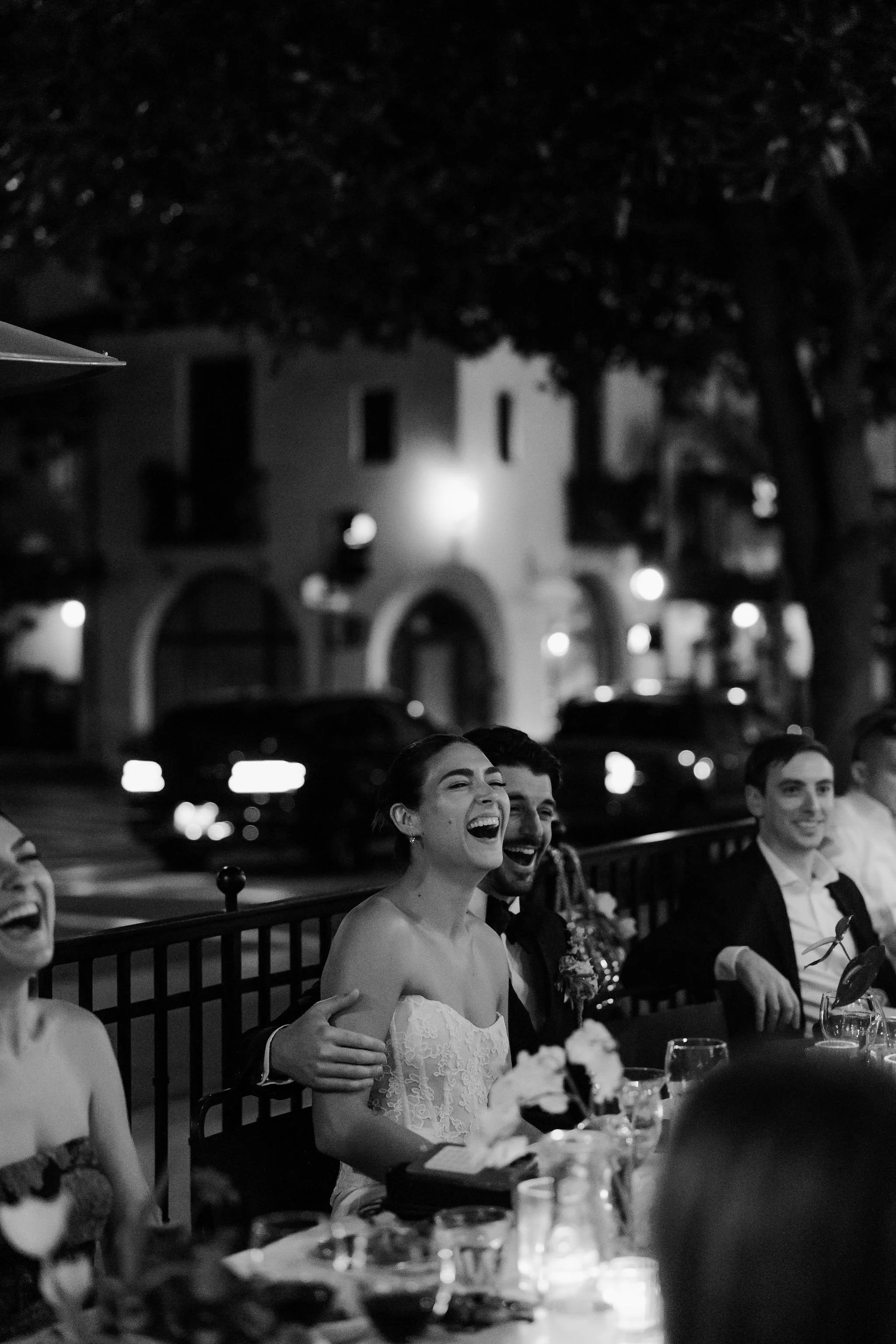 People sitting at a dinner table, dressed formally, smiling and laughing during a celebration, with a woman in a white dress at the center.