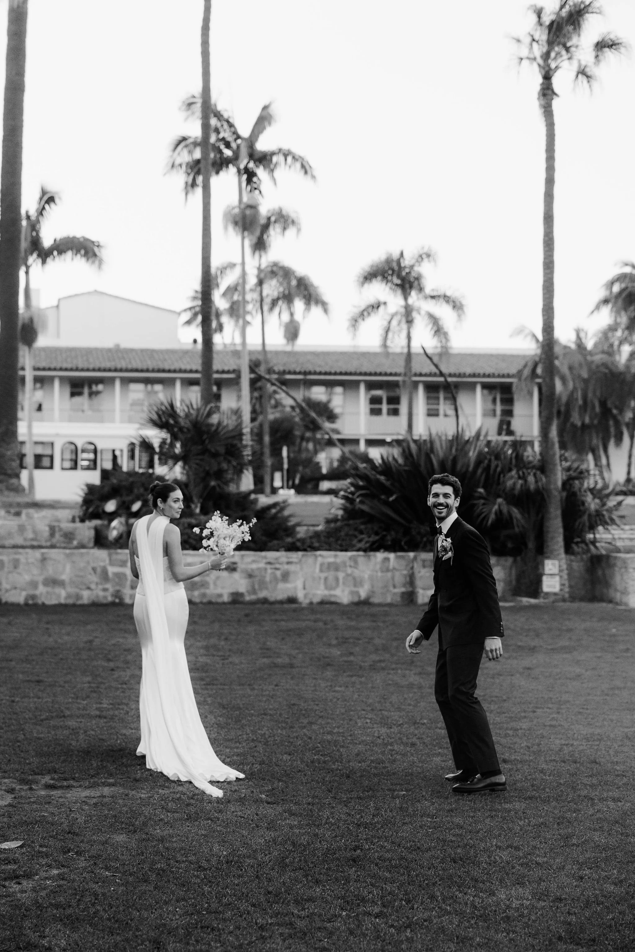 A black and white photo of a bride and groom outdoors. The bride is in a long, white gown holding a bouquet, looking at the groom. The groom is in a black suit, smiling, with palm trees and a building in the background.