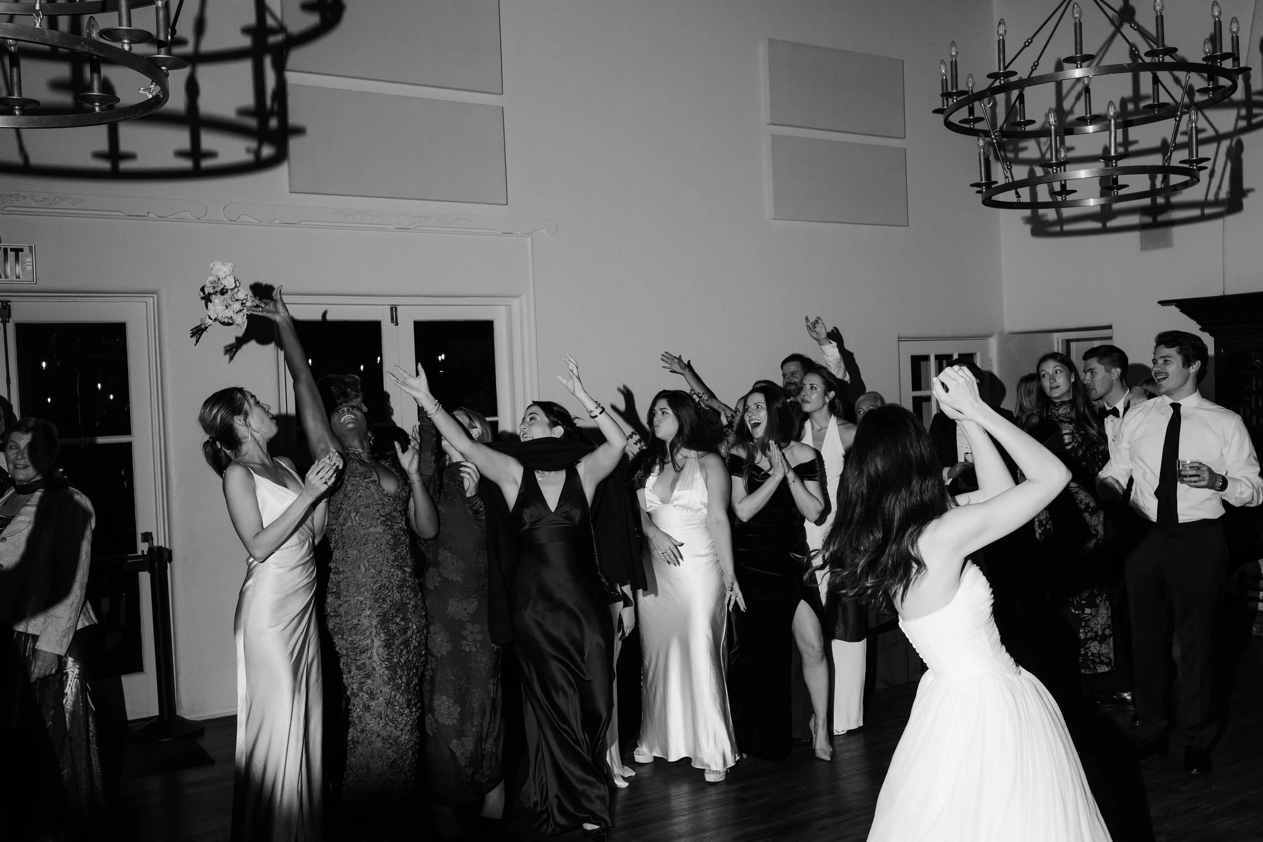 A black-and-white photo of a wedding reception with guests dancing and celebrating inside a decorated hall.