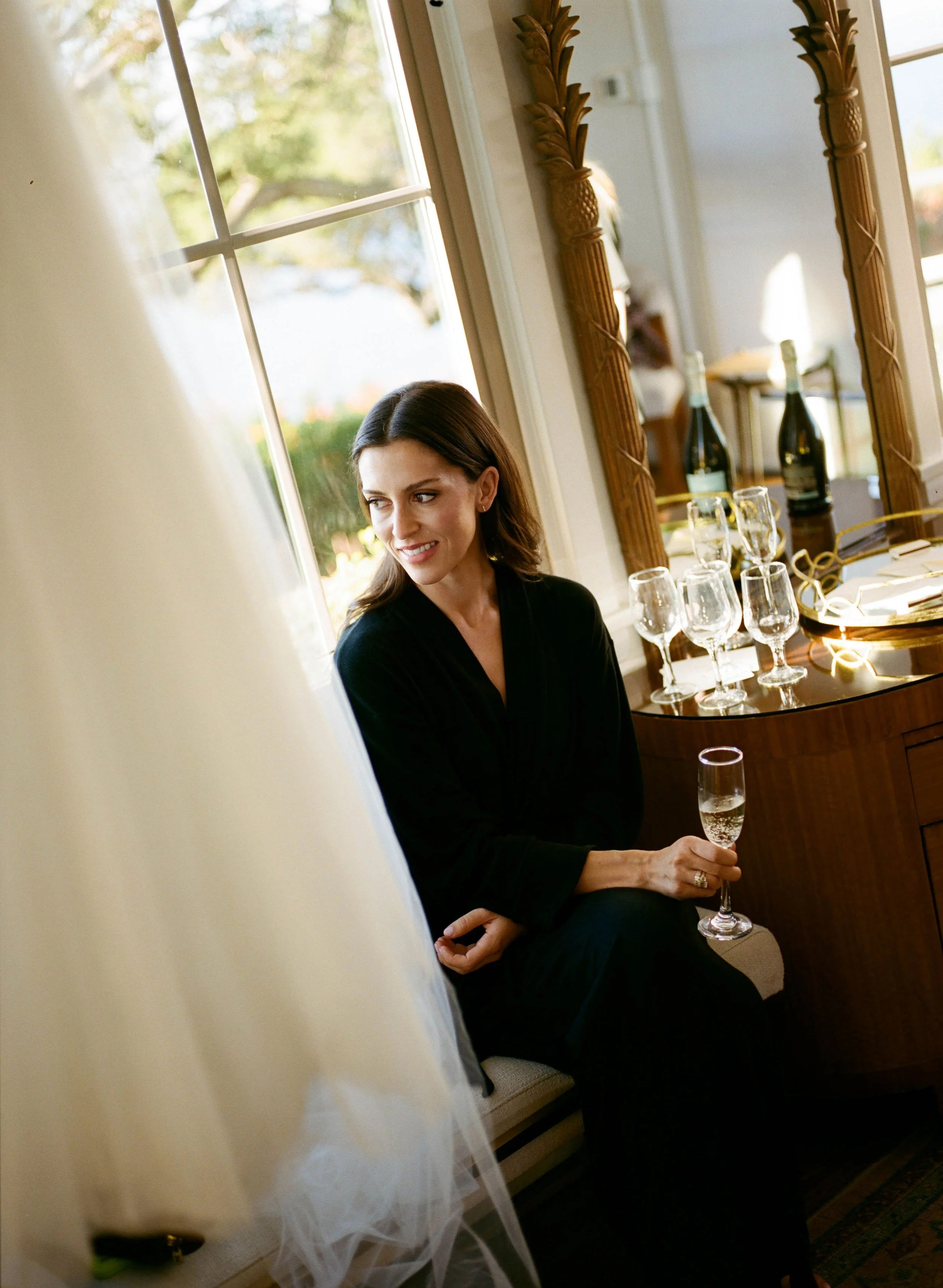 A woman with brown hair and light skin sitting indoors near a window, holding a flute of champagne, with a large mirror and decorated table with champagne bottles and glasses behind her.