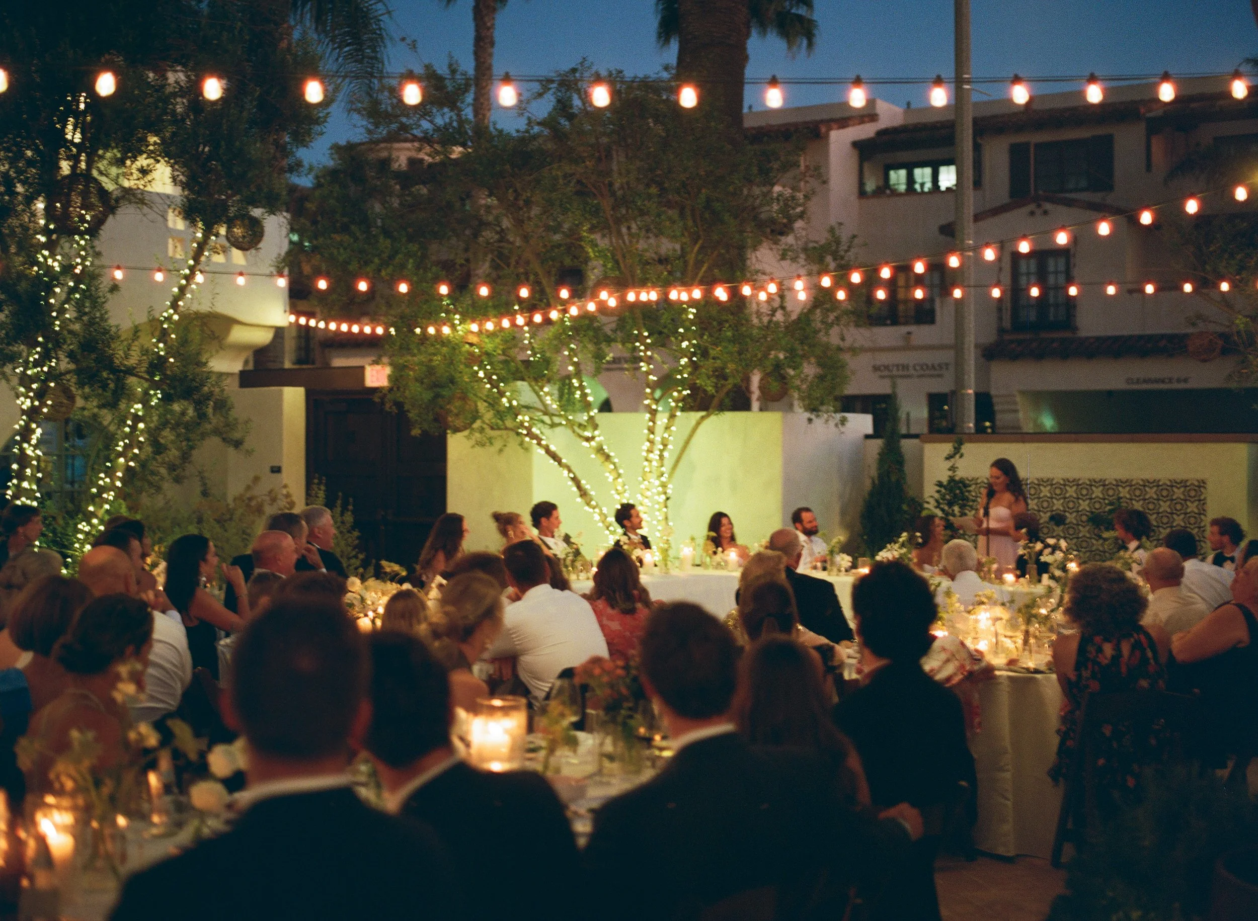 People seated at long tables with candles and floral centerpieces at an outdoor evening event under string lights, with buildings and trees in the background.