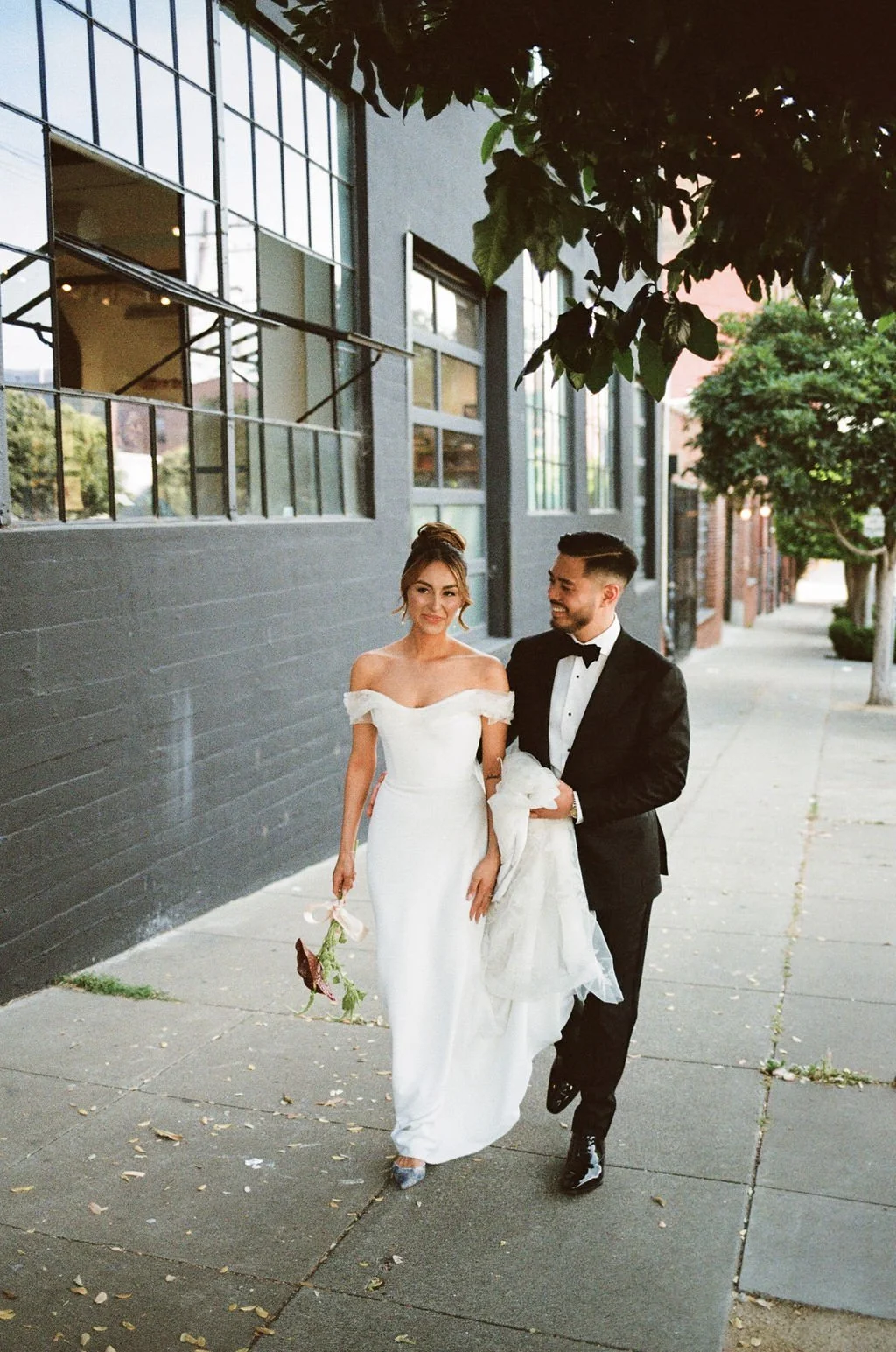 A bride and groom walking on a sidewalk, with the bride holding a bouquet, in an urban setting with trees and buildings in the background.