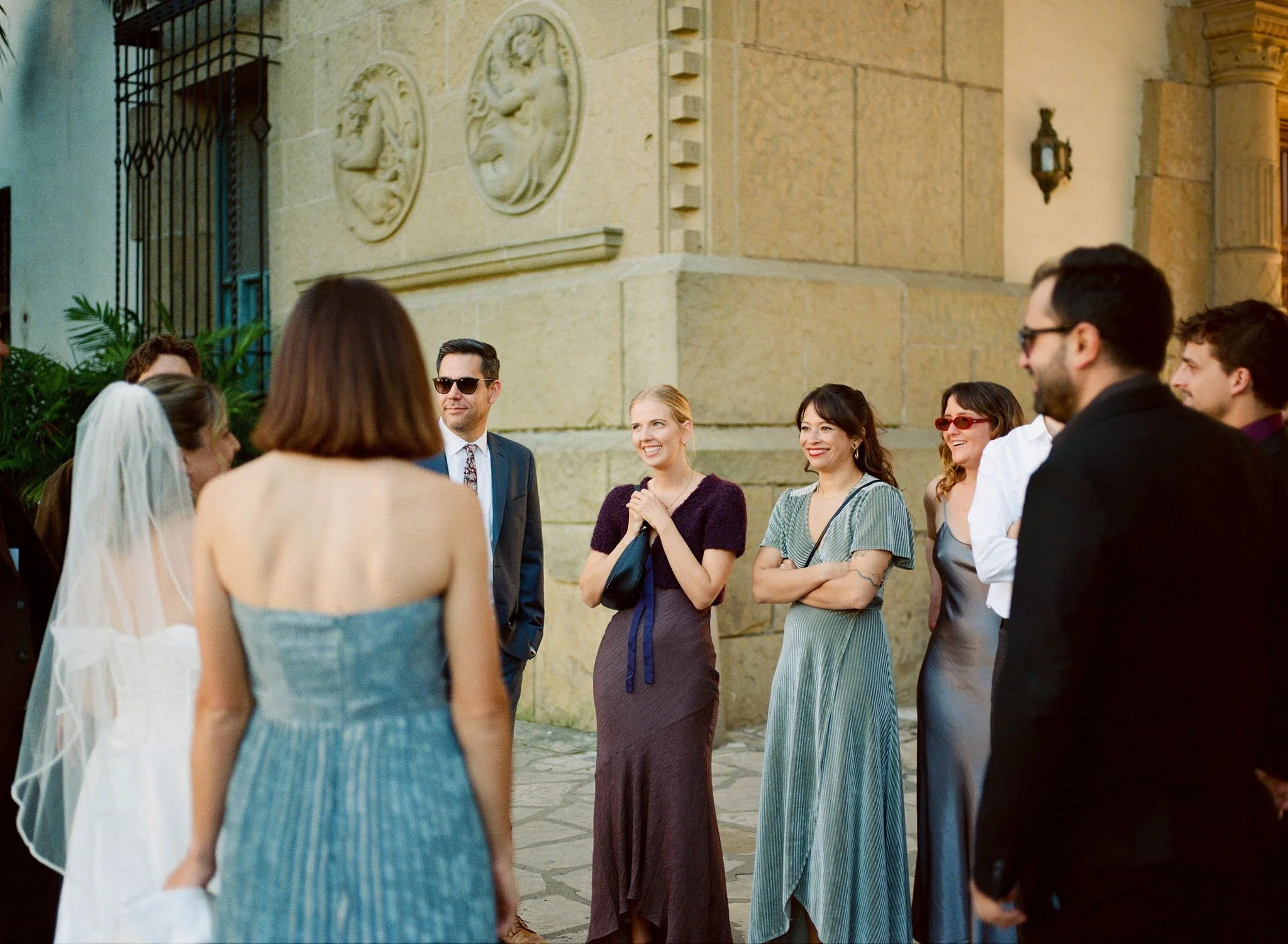 A group of people at a wedding or formal event standing outside near a stone building, engaging in conversation. The focus is on a smiling woman in a purple dress and a man with sunglasses in a suit.