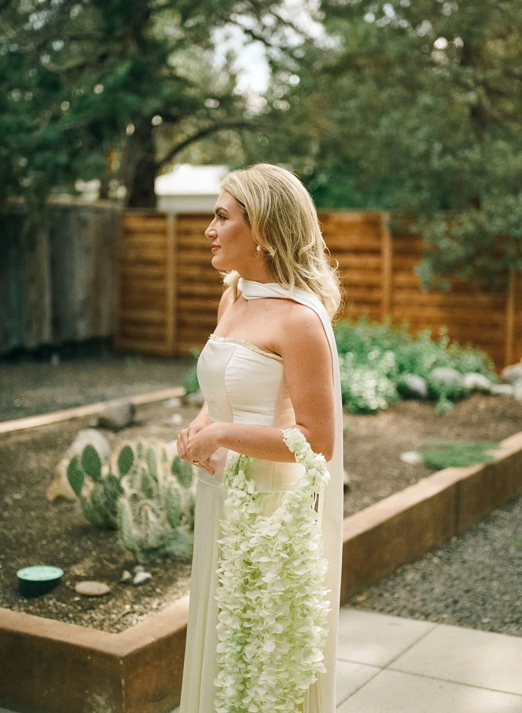A woman in a cream-colored dress holding a large white floral bouquet, standing outdoors with trees and a bamboo fence in the background.
