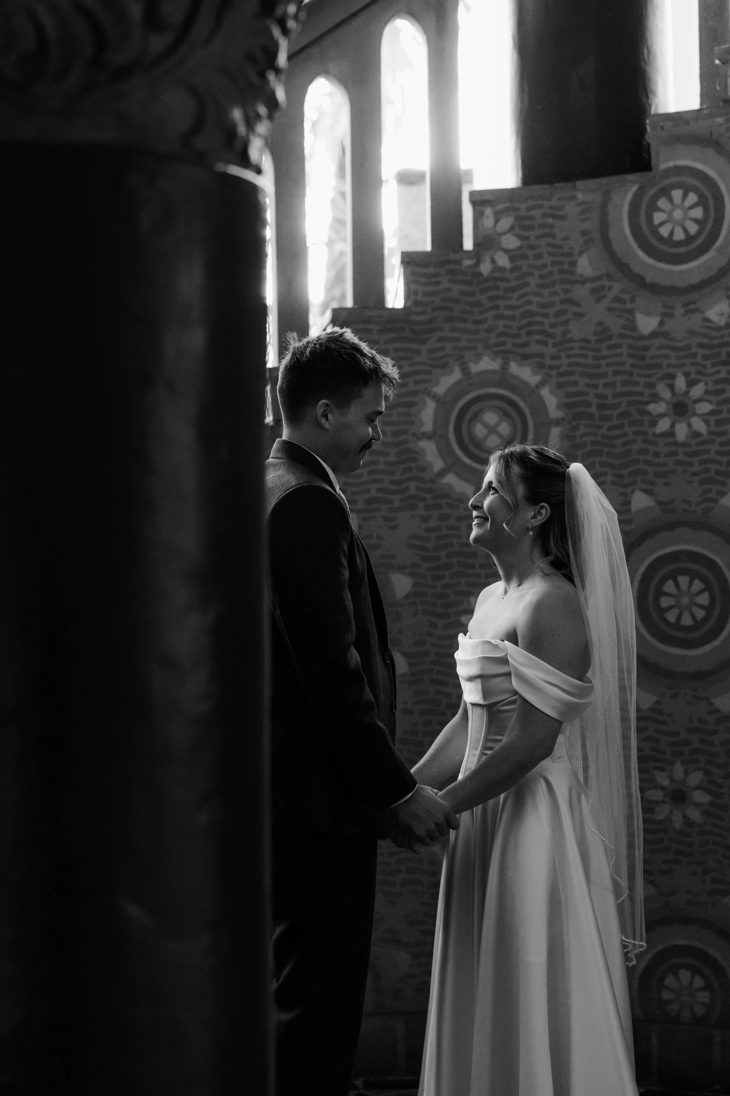 A black and white photo of a couple standing inside a church, holding hands and gazing at each other, with a stained glass window in the background.