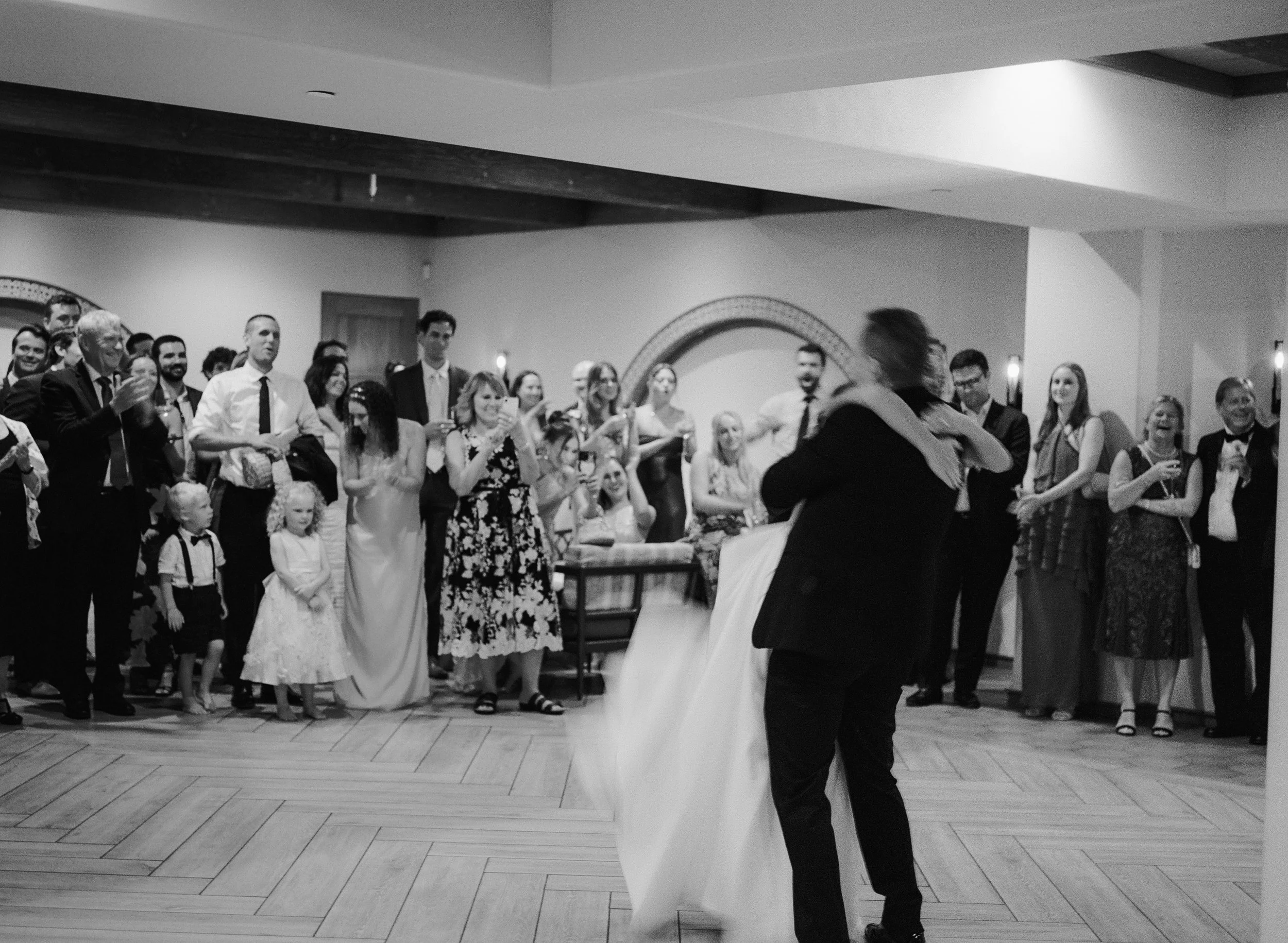 A group of people gathered indoors, watching a person in formal attire dancing or performing, with some taking photos and others smiling and clapping.