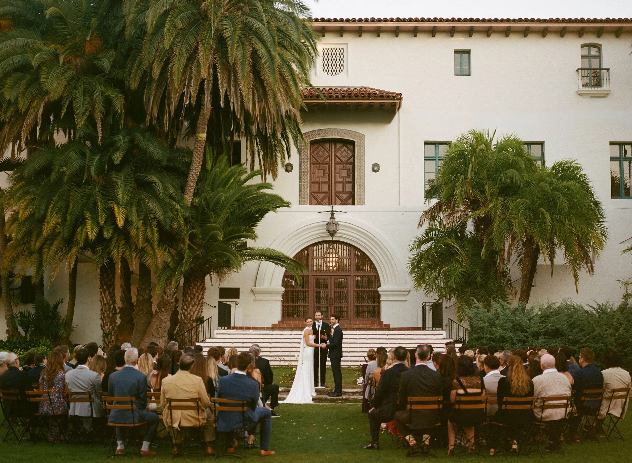 A wedding ceremony taking place outdoors at a historic building with white walls, red tiled roof, and numerous green palm trees surrounding the scene. The bride and groom stand before an officiant, with guests seated on chairs watching. The setting i