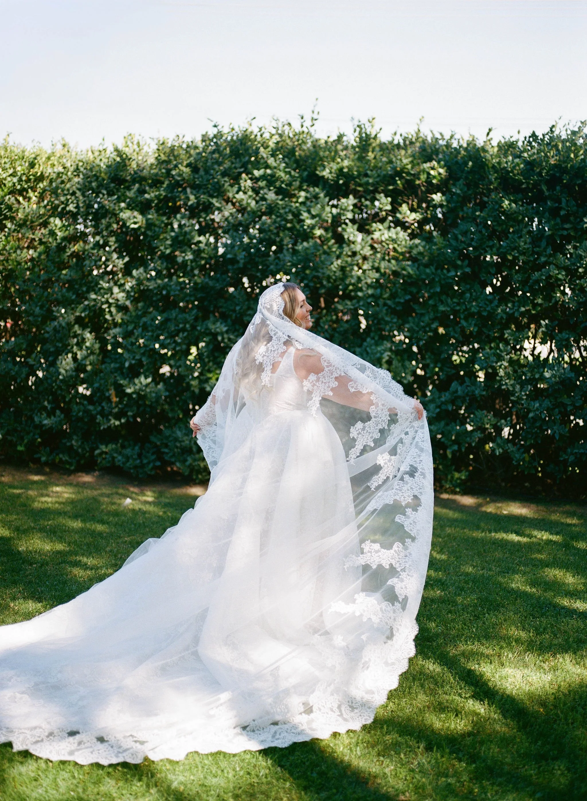 A bride in a wedding dress with a lace veil holding the edges of her dress outdoors in front of a large green bush, with sunlight casting shadows on the grass.