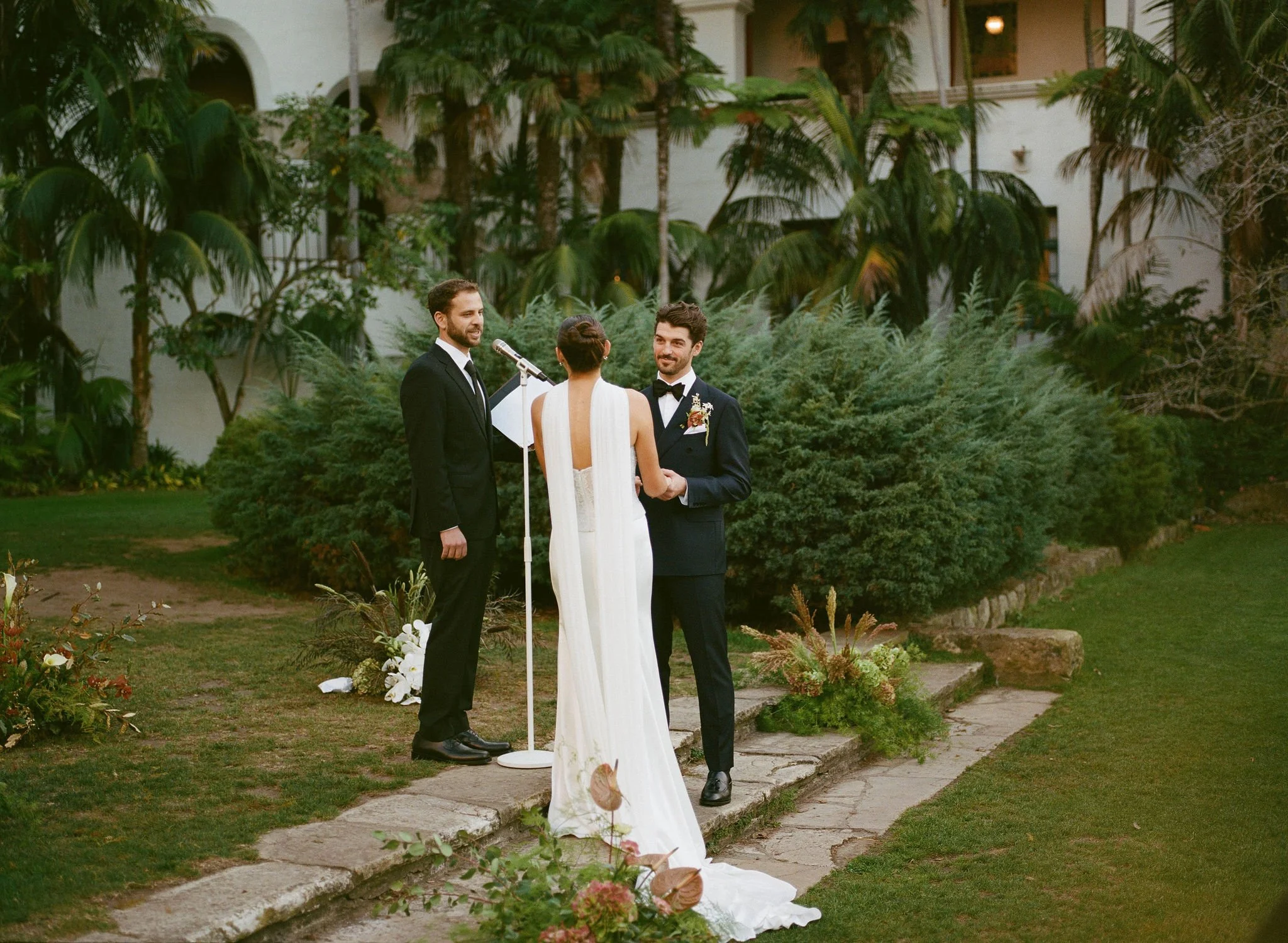 A couple getting married outdoors with an officiant during sunset, surrounded by lush greenery and a large building in the background.