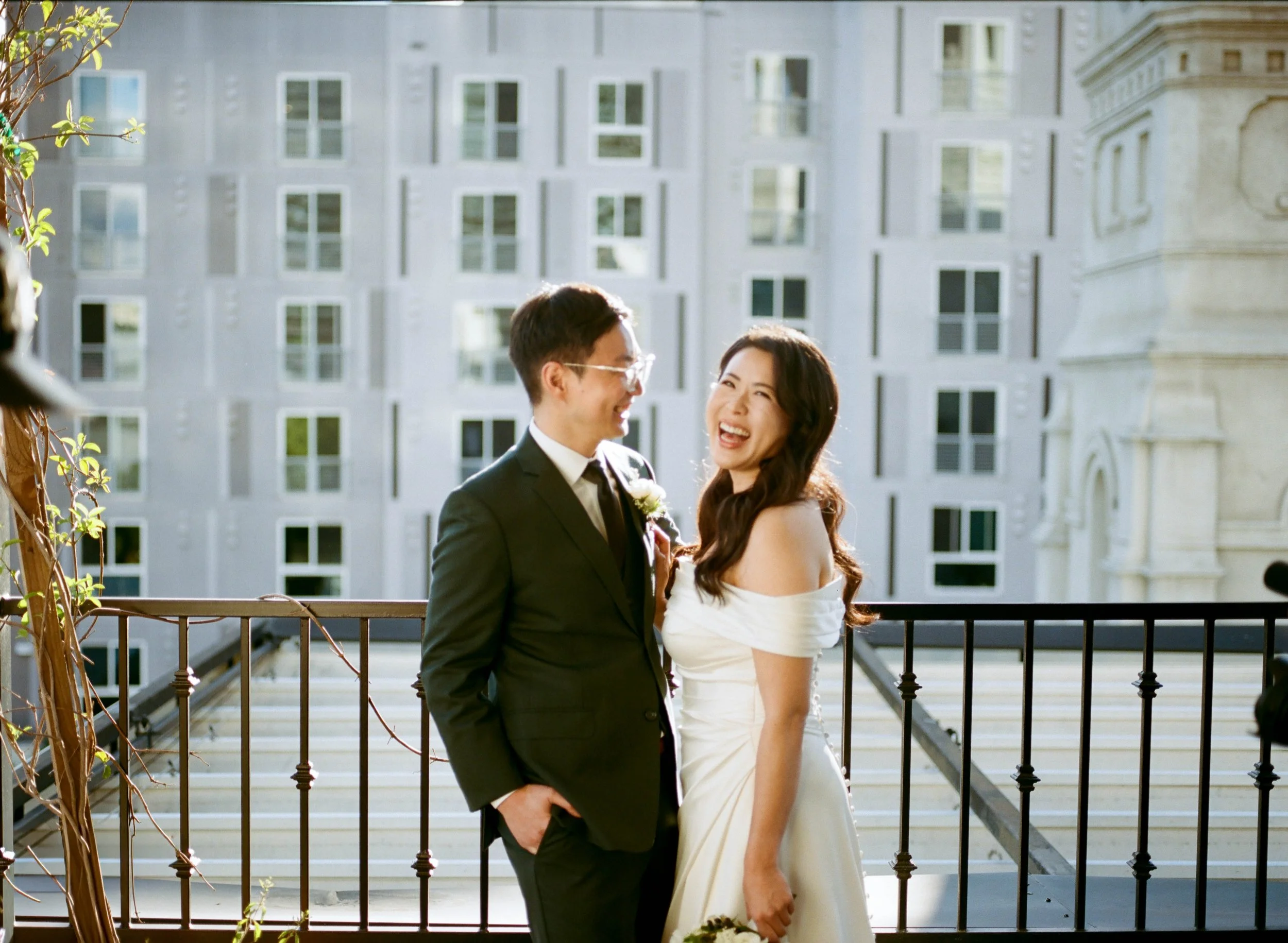A smiling couple, dressed in formal attire, standing on a balcony in front of a large white building, possibly during a wedding or special event.
