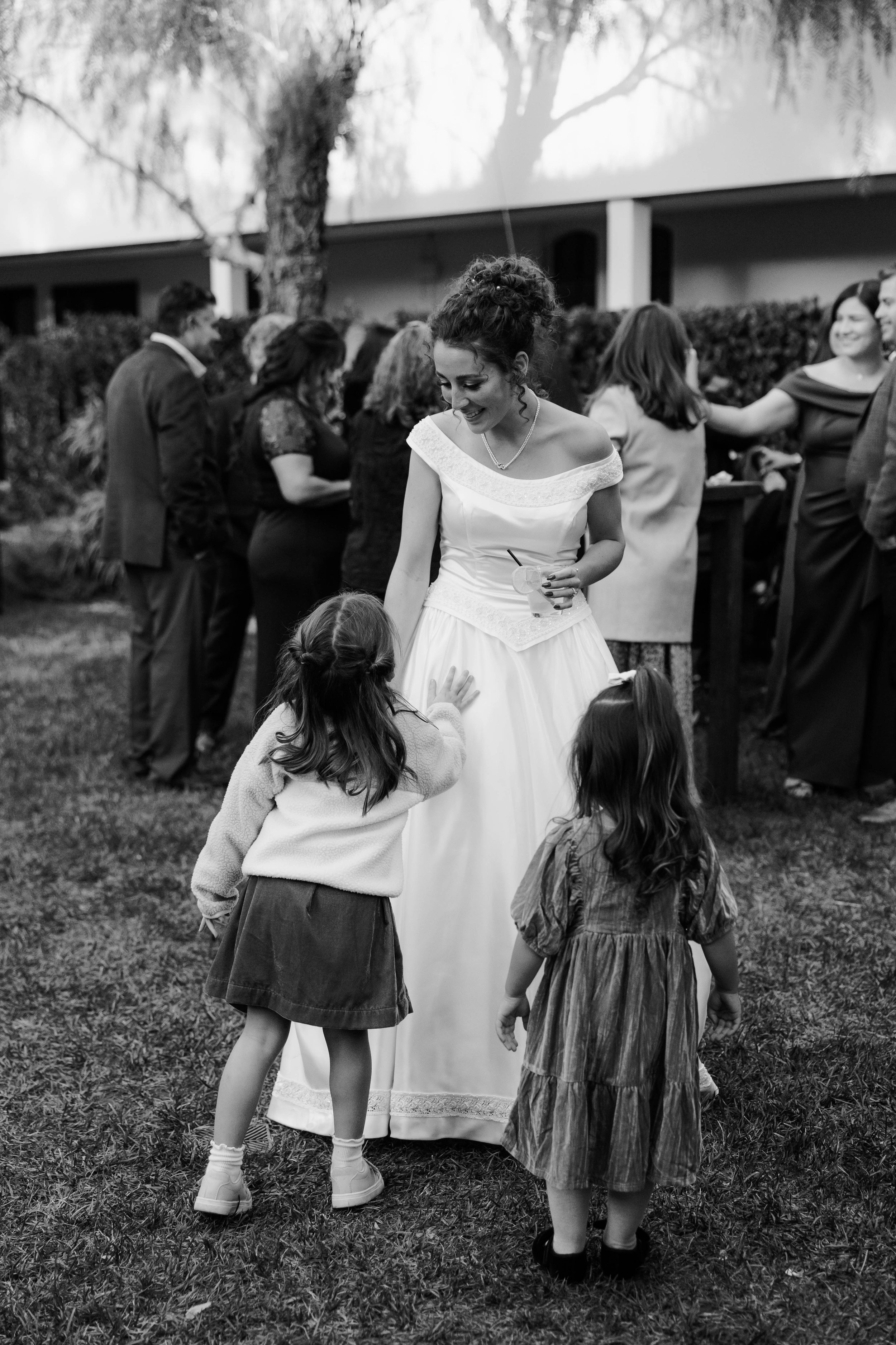 A black and white photo of a woman in a wedding dress smiling and talking to two young girls at an outdoor gathering with other people in the background.