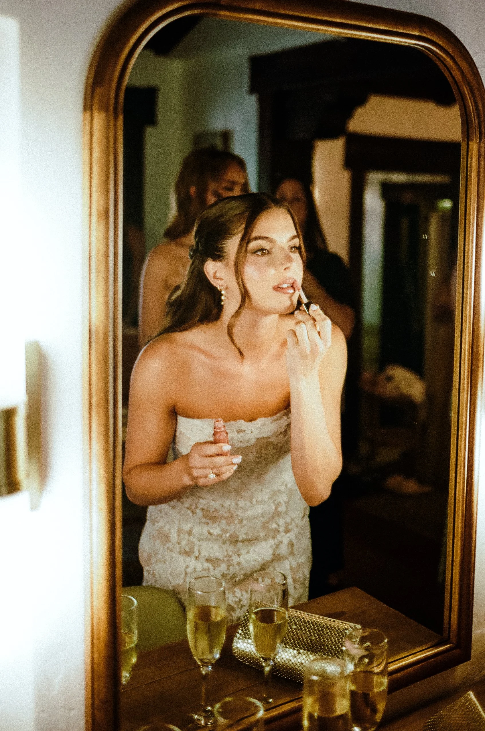 A woman in a lace strapless dress is applying lipstick while looking at herself in a mirror. She is holding a lipstick in her right hand, and her reflection shows her in a room with other women in the background. The table in front of the mirror has 