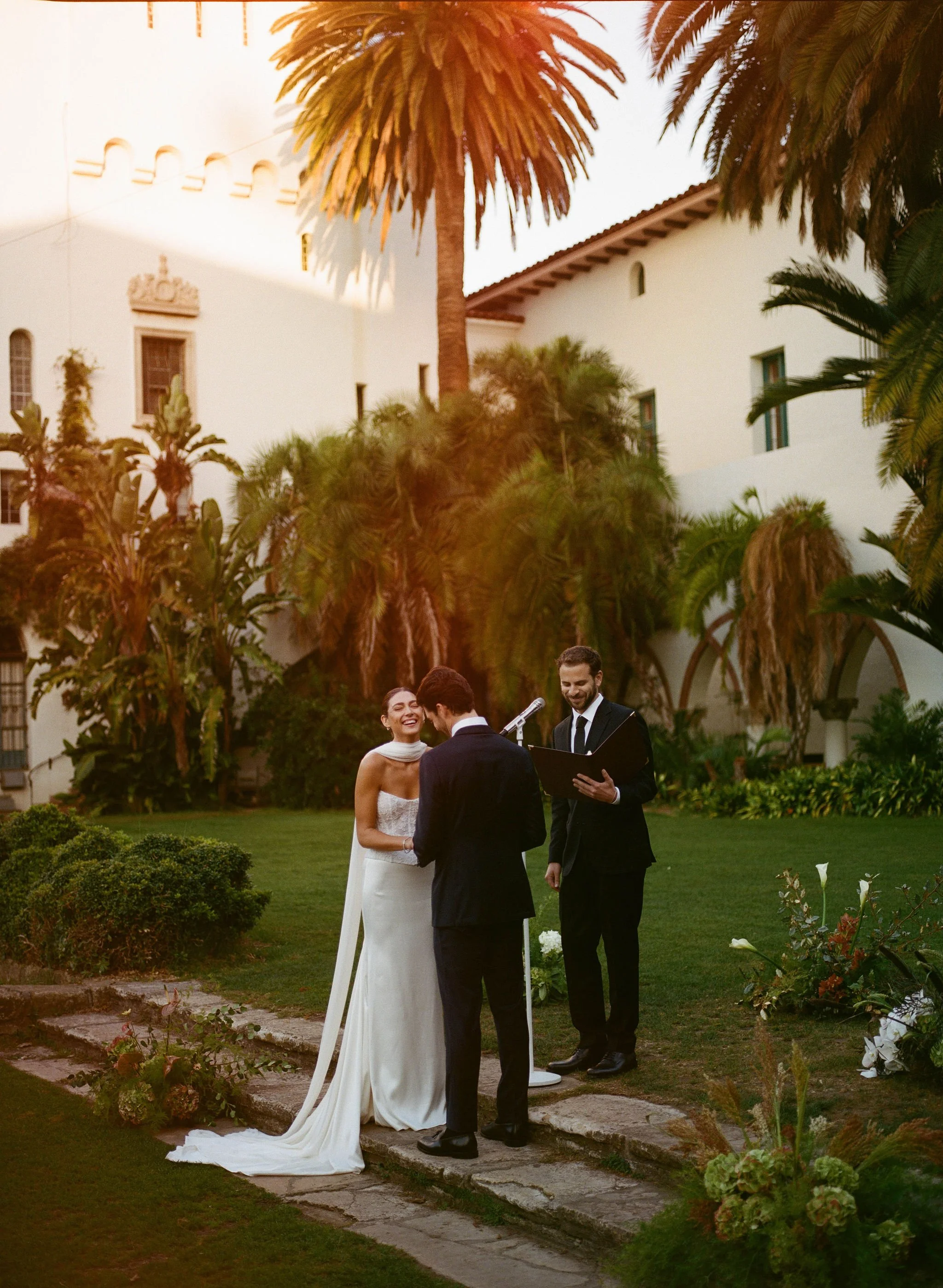 A couple getting married outdoors, with a man and a woman standing together in front of an officiant, surrounded by trees and a white building.