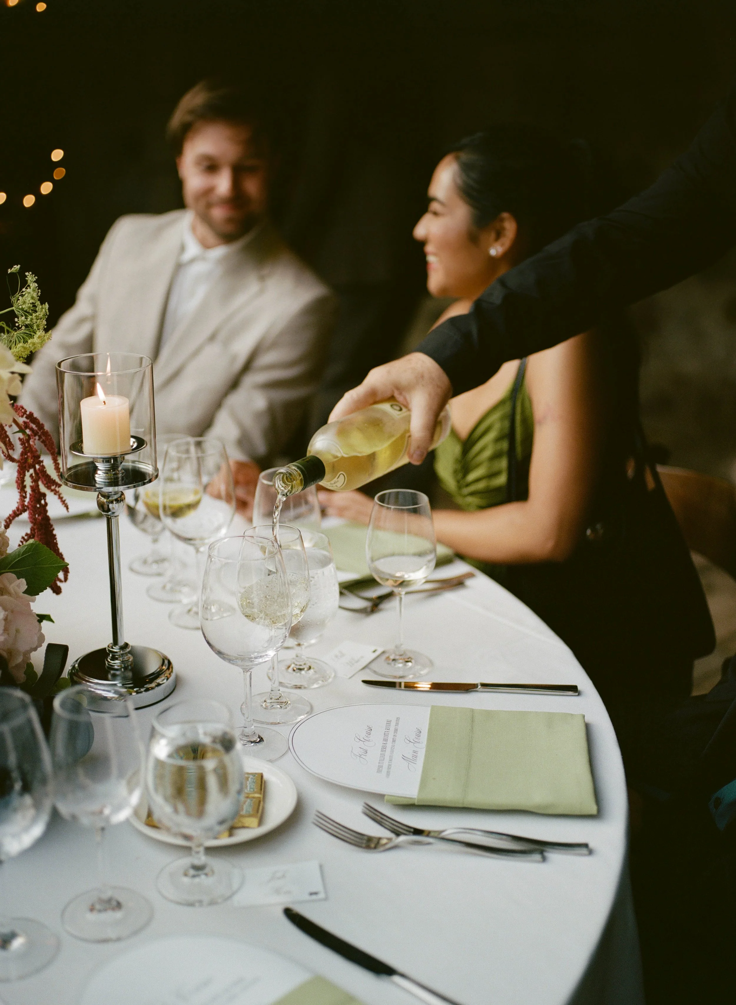 A romantic dinner scene with a man in a light-colored suit and a woman in a green dress, smiling and enjoying a moment. A person pouring white wine into glasses at a neatly set table decorated with flowers and a candle.