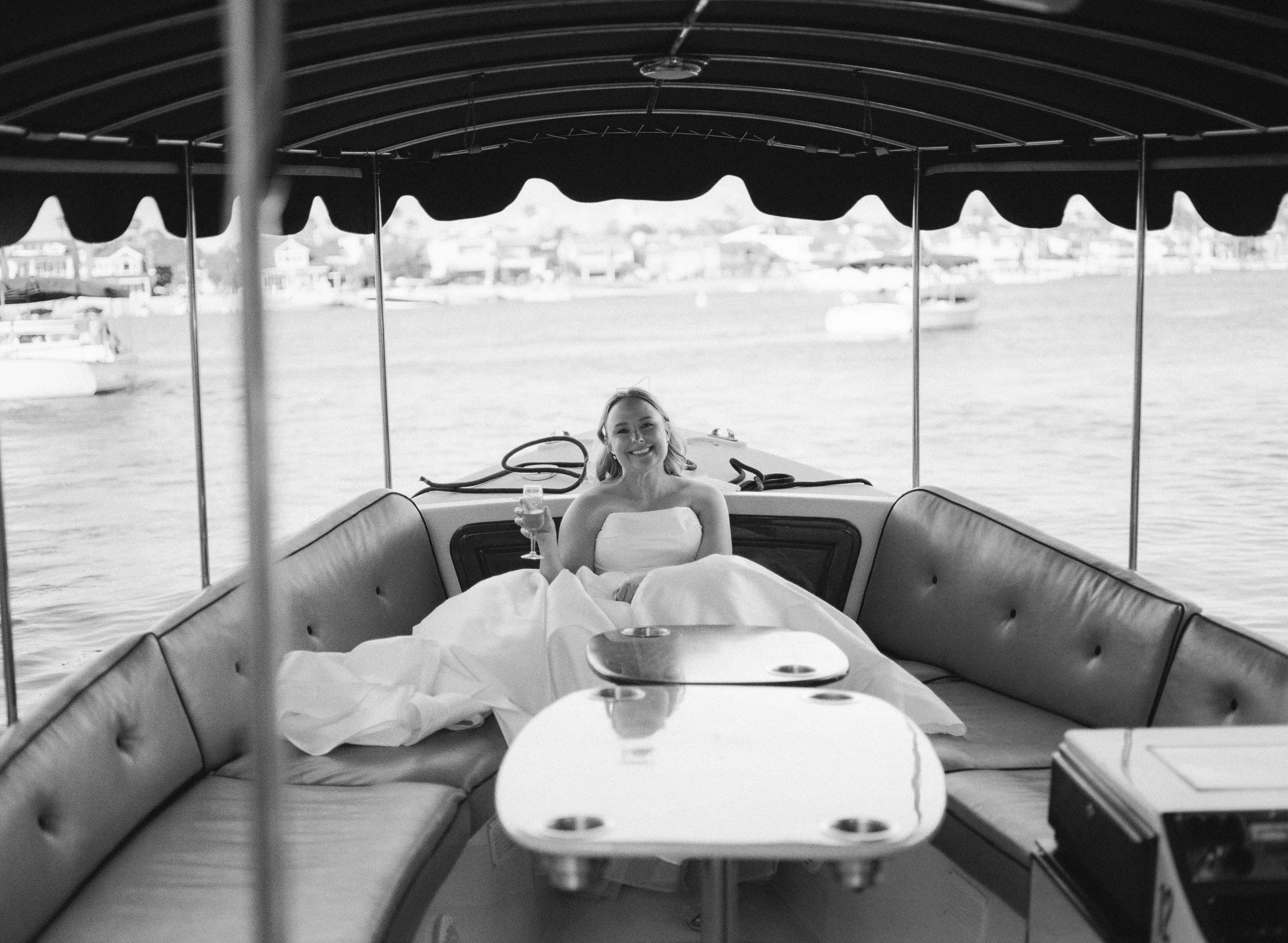 A woman in a swimsuit sitting on a boat holding a glass of wine, with water and city buildings in the background.