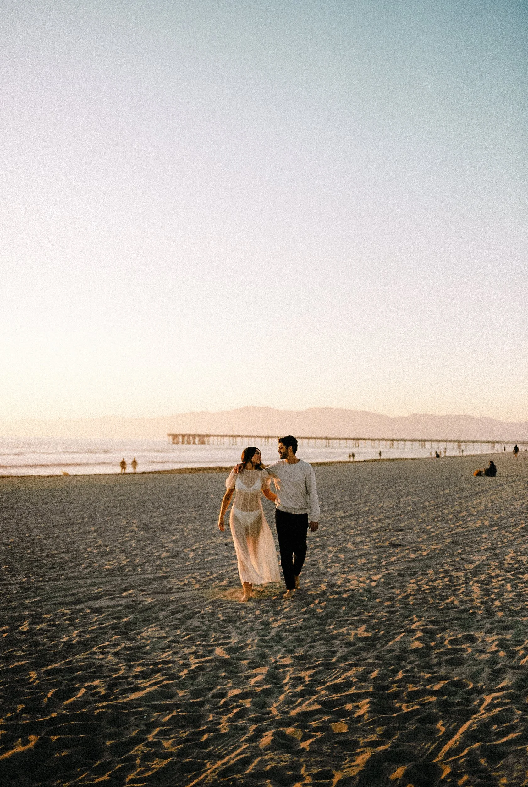 A couple walking on the beach at sunset. The woman is wearing a sheer white dress, and the man is dressed in a light gray shirt and dark pants. The scene is peaceful, with a pier visible in the background and a few people scattered along the shorelin