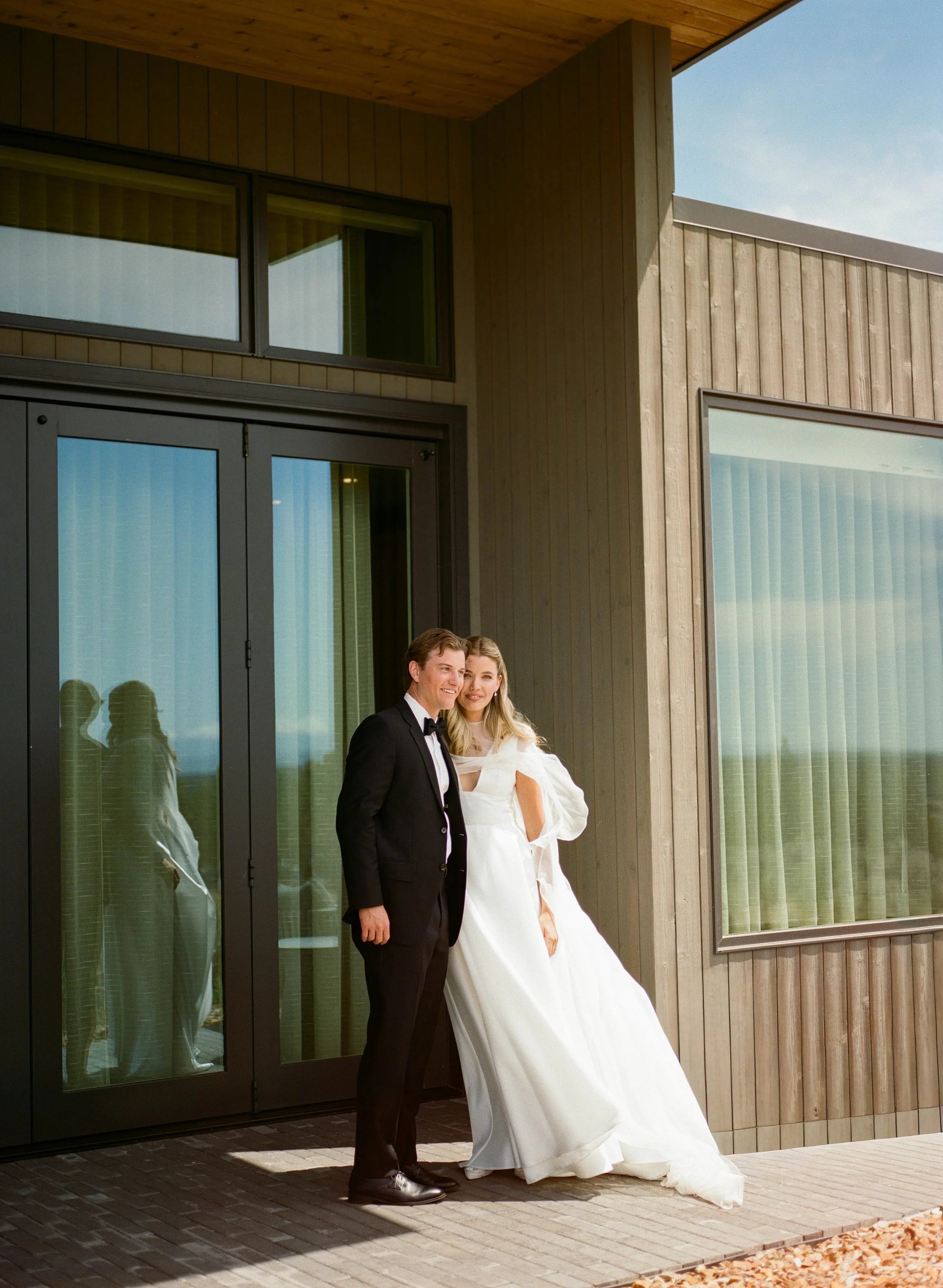 A couple dressed in wedding attire standing outside a modern building with large windows and wooden siding.