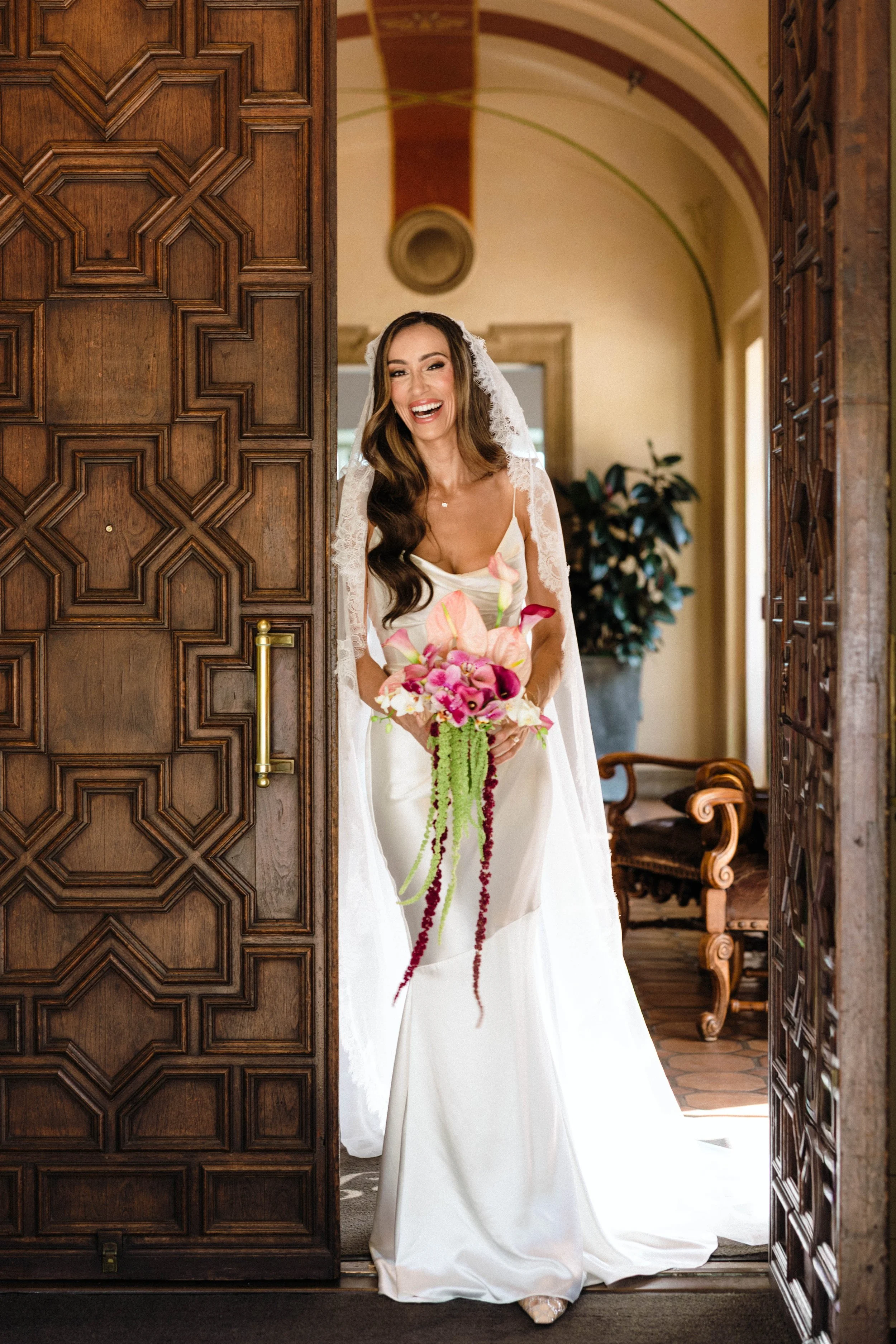 A woman wearing a white wedding dress and veil holding a pink and white flower bouquet, smiling and standing in a doorway between two wooden doors inside a room with high ceilings and a potted plant in the background.