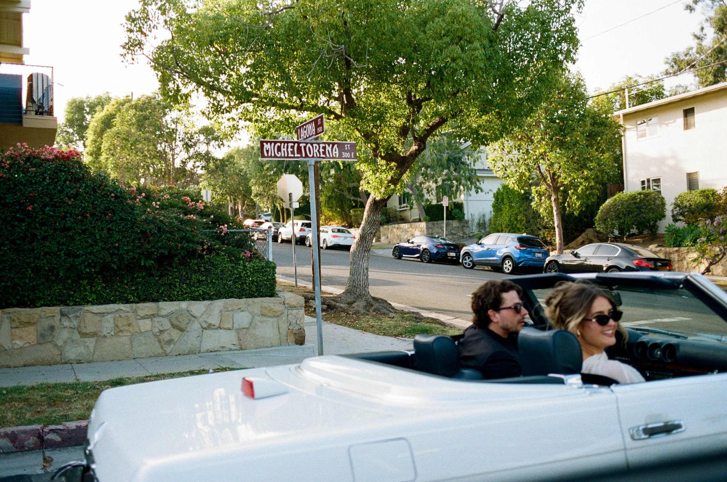 A neighborhood street scene with street signs for Laguna Street and Michel Torrena Street, parked cars along the curb, trees, and residential buildings in the background. Two people are in a convertible car in the foreground, smiling and wearing sung