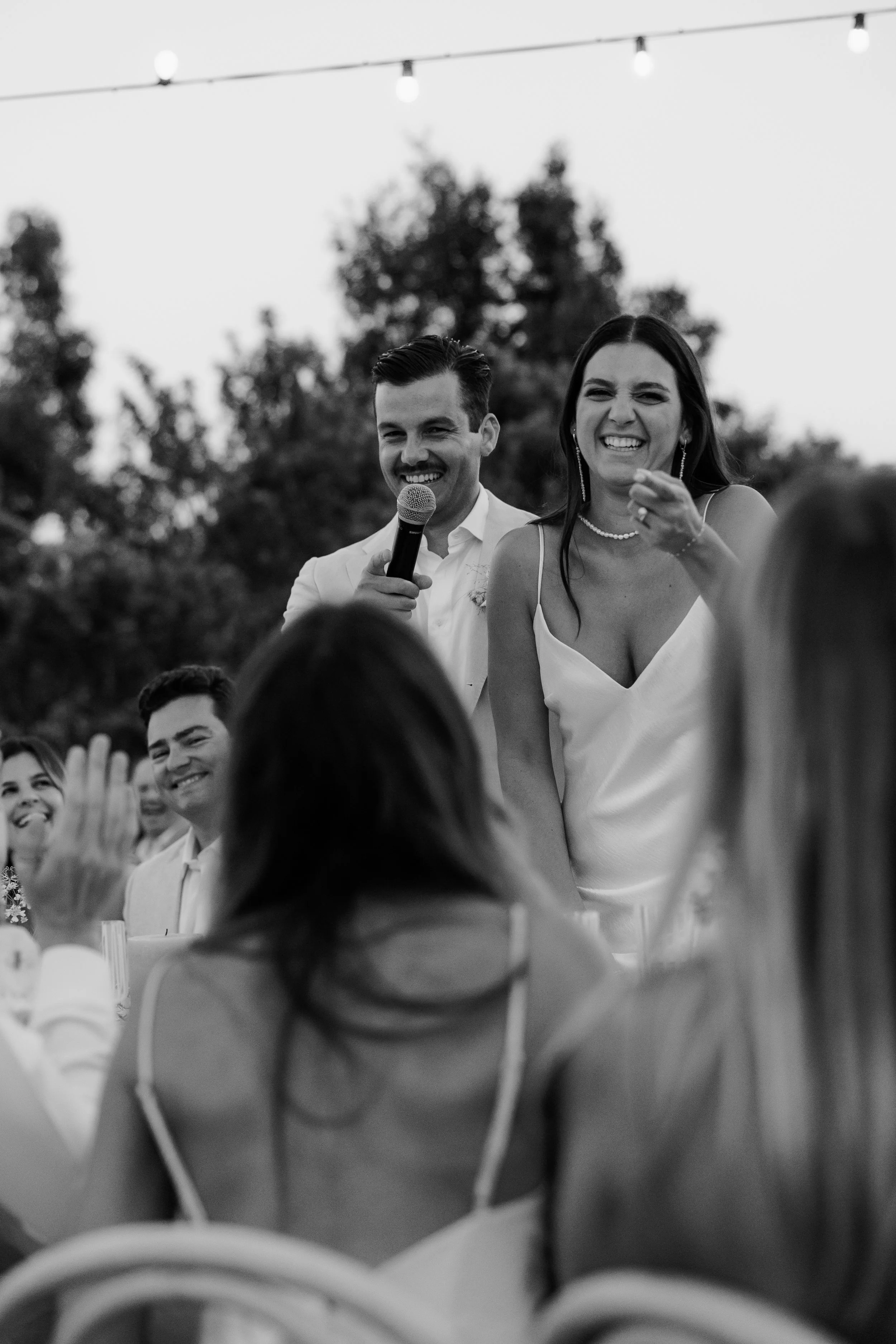 Black and white photo of a woman in a dress and a man holding a microphone, both smiling at a gathering outdoors with string lights and trees in the background.