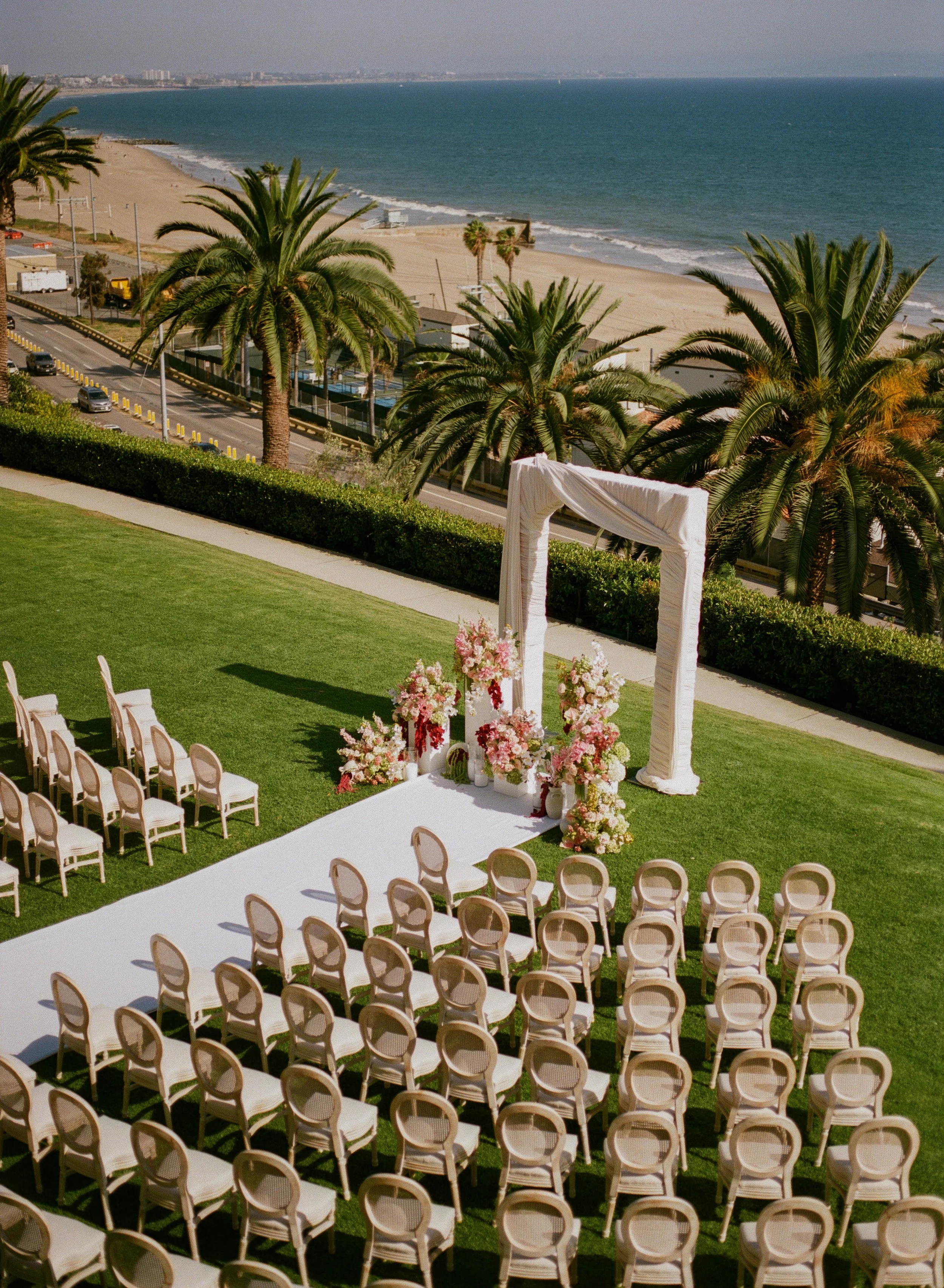 Outdoor wedding setup on a grassy area overlooking the beach, with rows of chairs, a floral arch, and ocean views in the background.