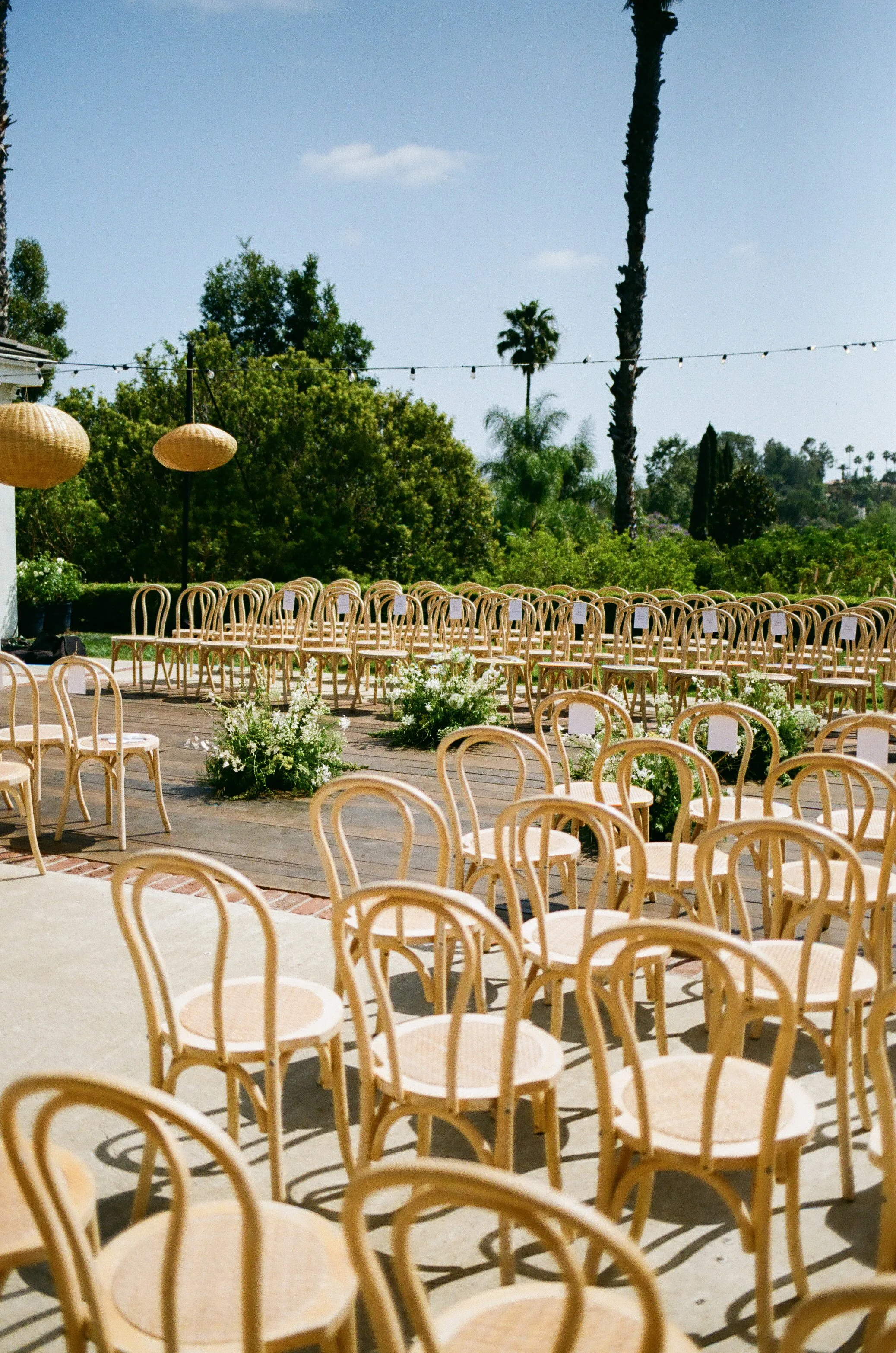 Outdoor wedding ceremony setup with rows of beige chairs, potted flowers, string lights, and tall palm trees under a clear blue sky.