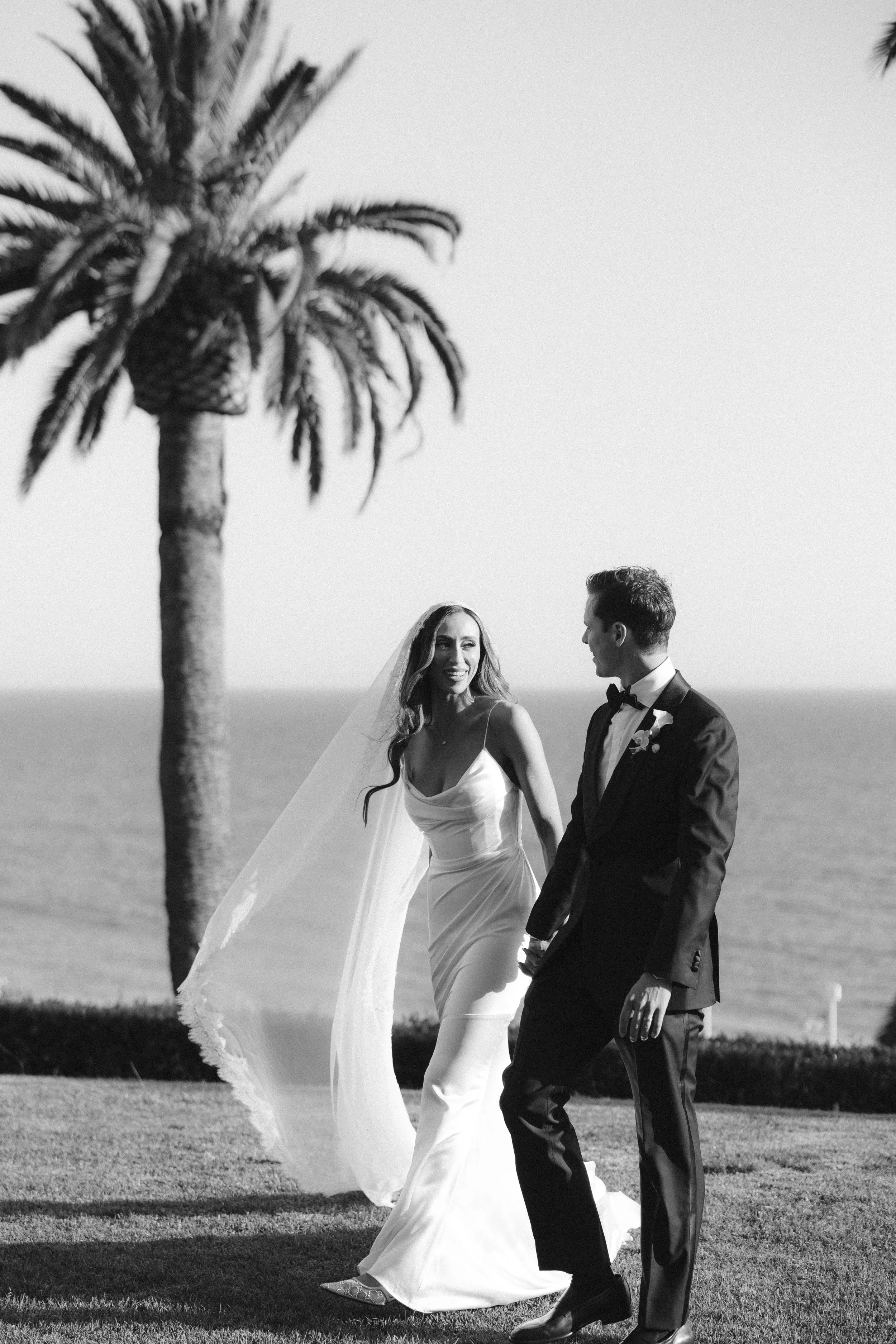 A black and white photo of a bride and groom walking outdoors near a palm tree, holding hands and smiling at each other.