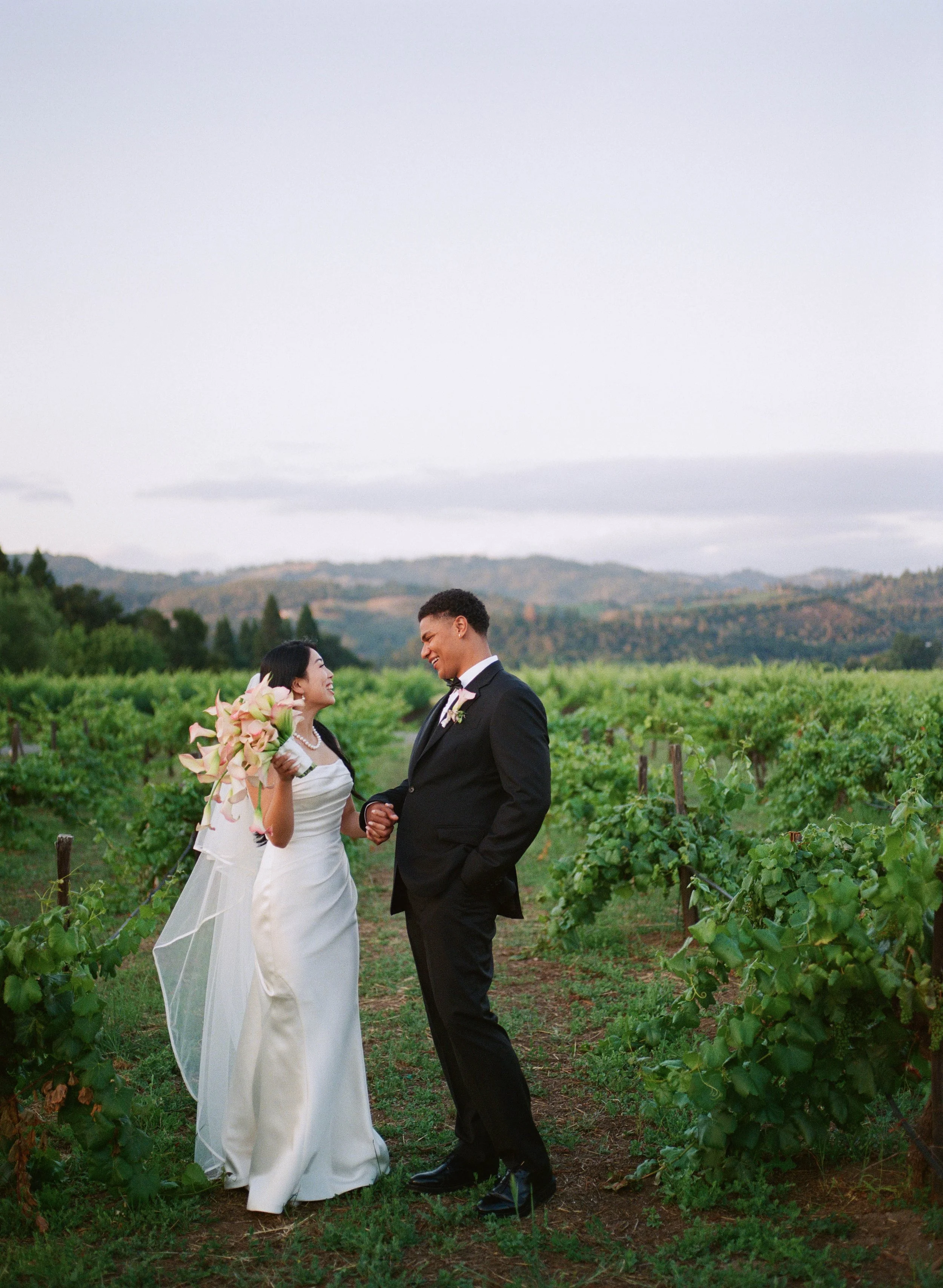 A bride and groom hold hands and smile at each other in a vineyard landscape during their wedding.