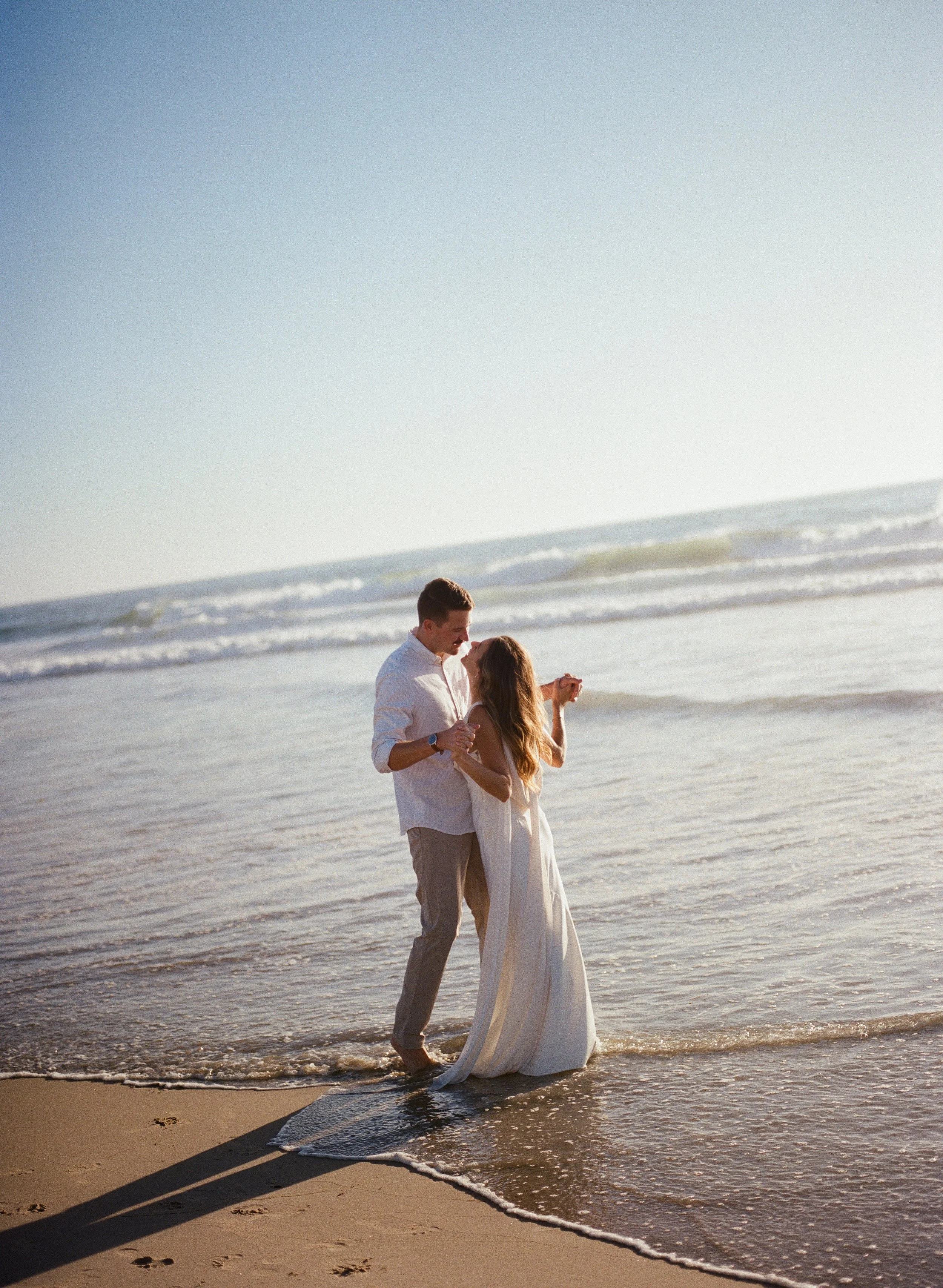 A couple is dancing on the beach at sunset, with the ocean waves in the background.