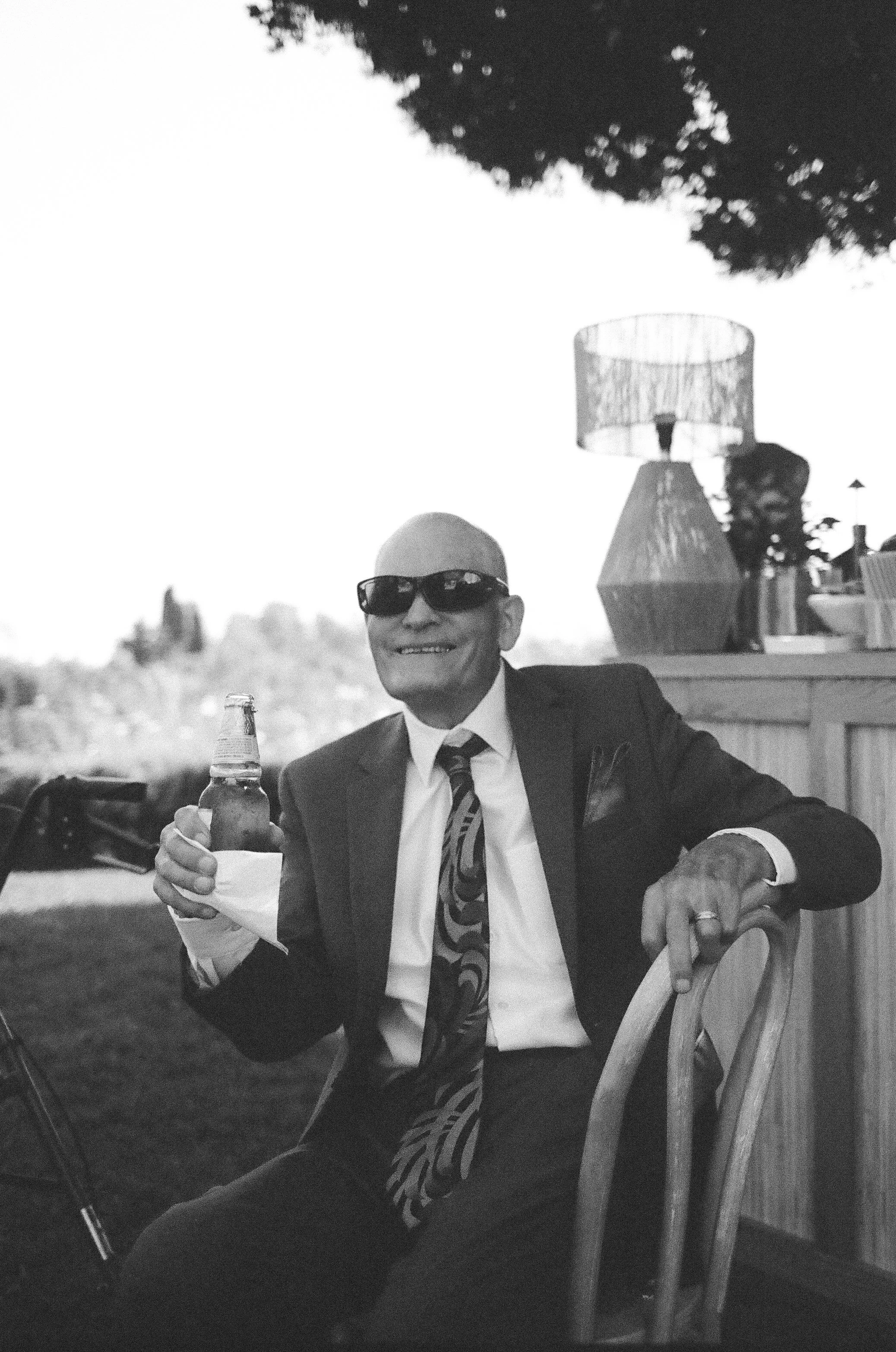 A man wearing a suit, tie, and sunglasses sitting at a table outdoors, holding a beer bottle, with a smile. There is a lamp and a bottle on the table. The background shows trees and buildings in the distance.