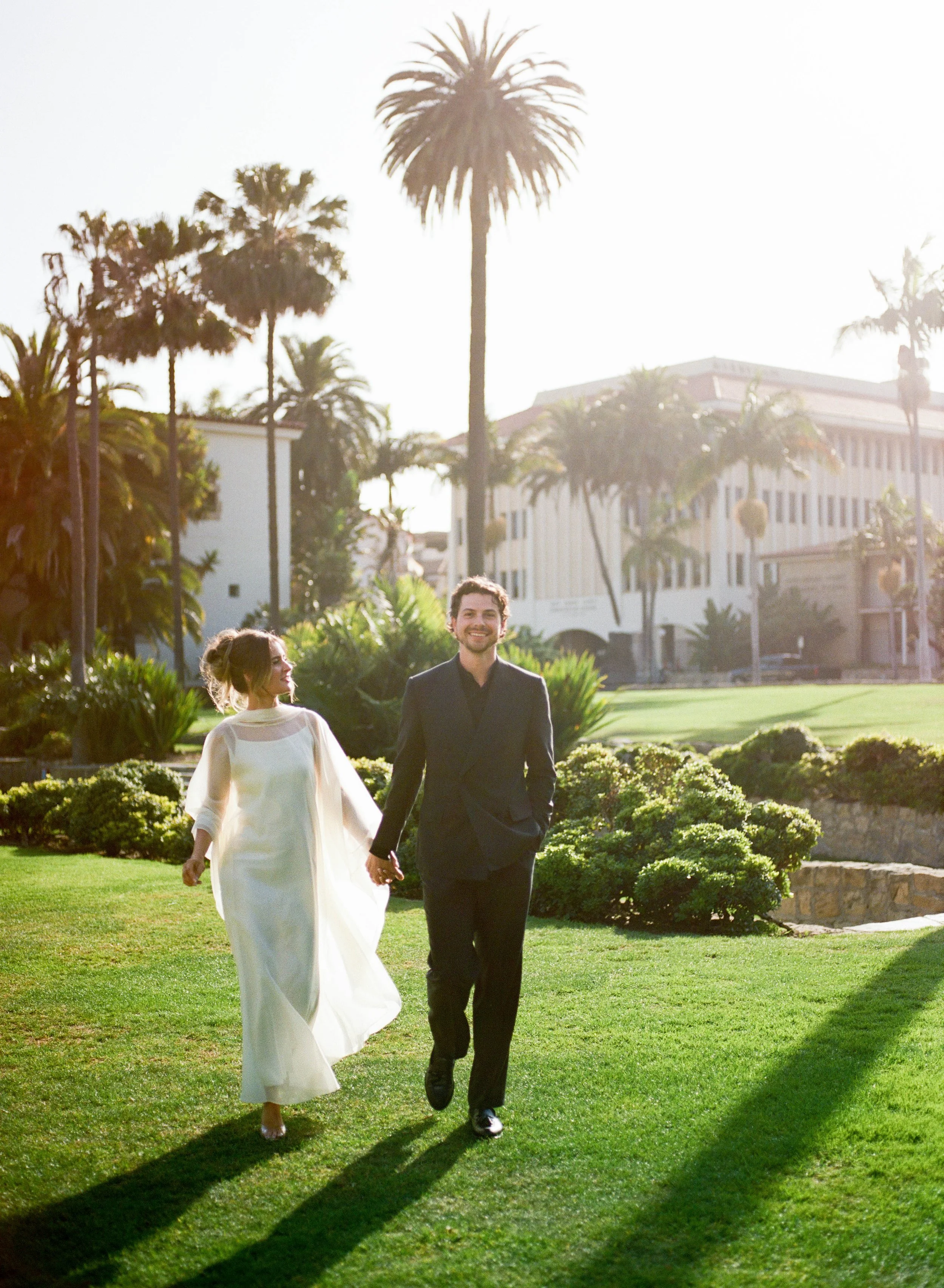 A smiling man in a black suit and a woman in a white dress walking hand in hand on a lush green lawn, with palm trees and a building in the background, under bright sunlight.