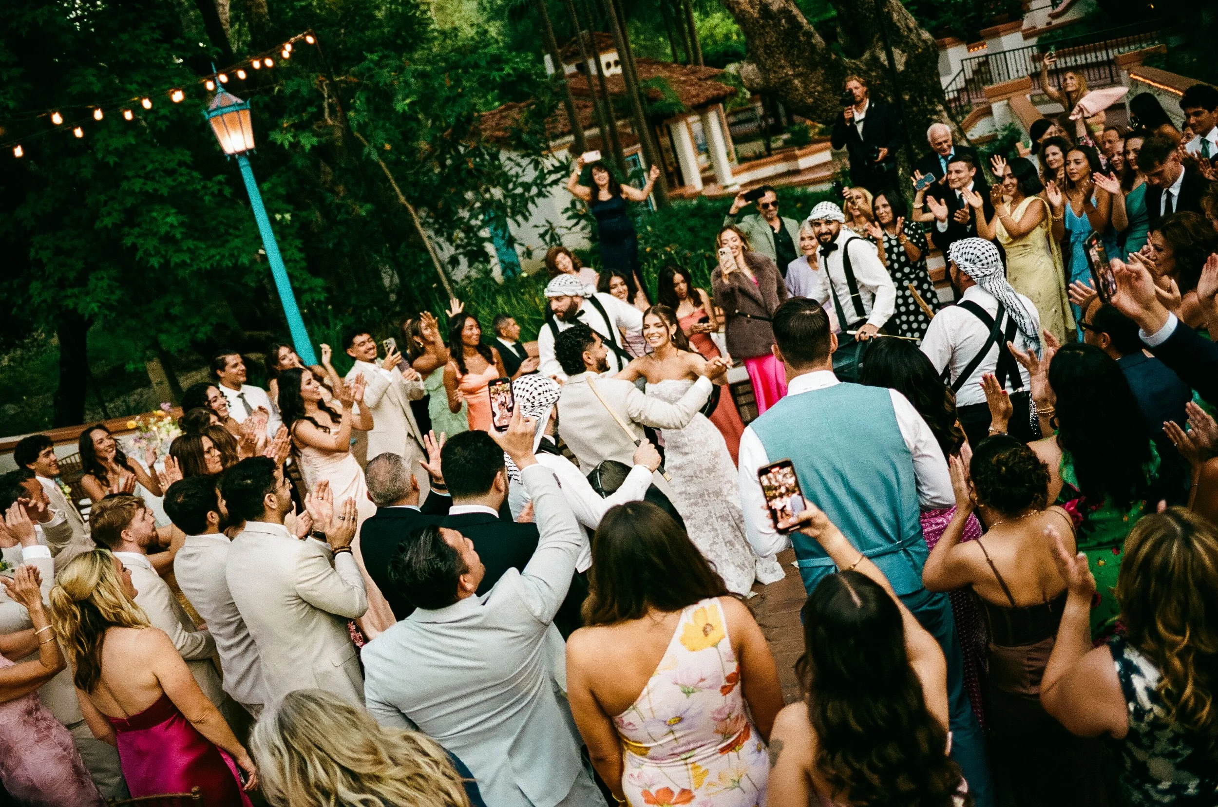 A large gathering of people at an outdoor wedding reception with a bride and groom dancing in the center. Guests are clapping, taking photos, and enjoying the event under string lights and surrounded by trees and greenery.