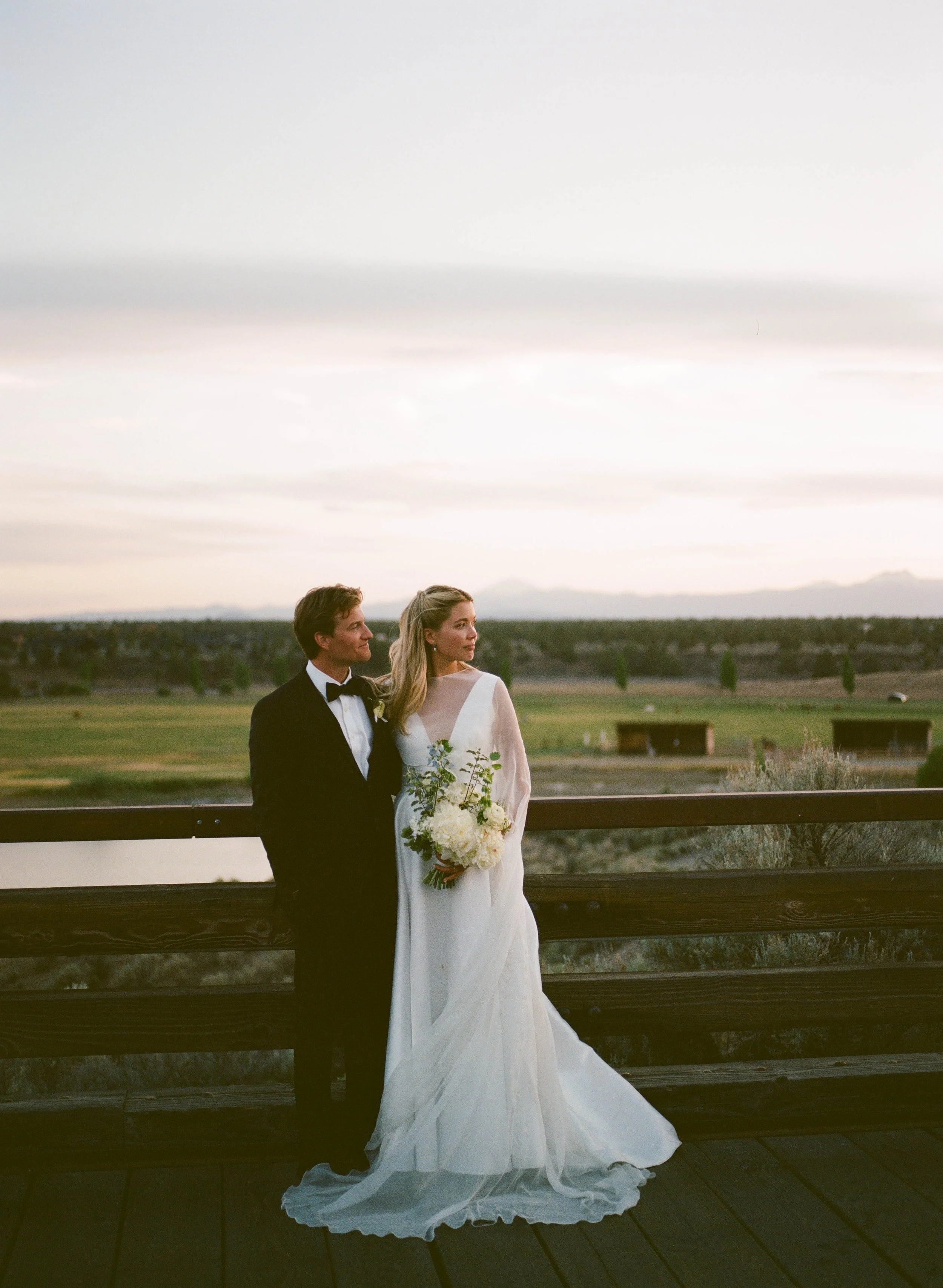 A bride and groom standing on a wooden bridge outdoors at sunset, with a scenic open field and cloudy sky in the background. The bride is holding a bouquet of white flowers and wearing a white wedding gown, while the groom is in a black tuxedo with a