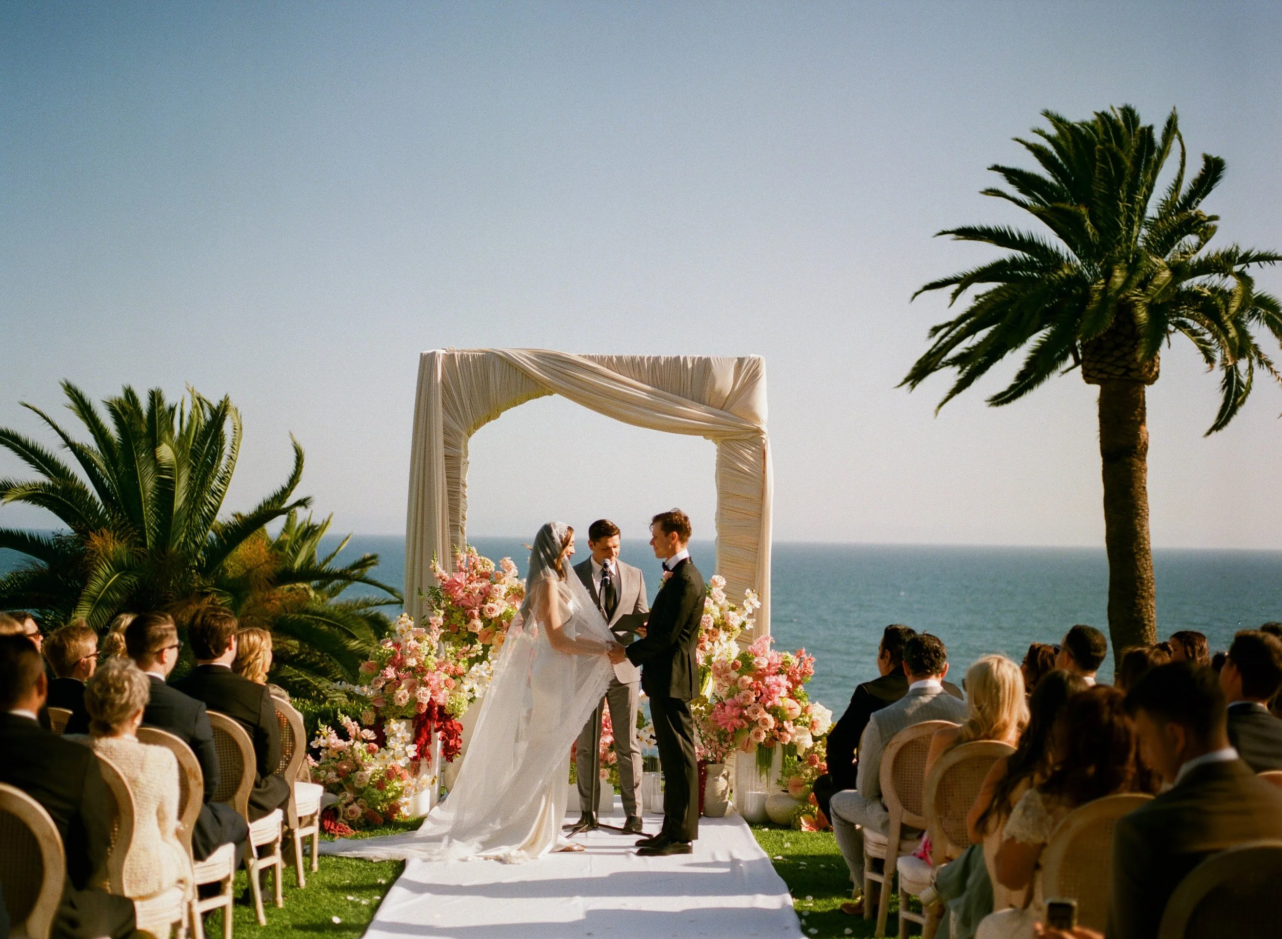 A wedding ceremony taking place outdoors with the ocean in the background. The bride and groom are exchanging vows under a decorated arch with flowers, surrounded by guests seated on either side. Tall palm trees frame the scene, and the sky is clear 