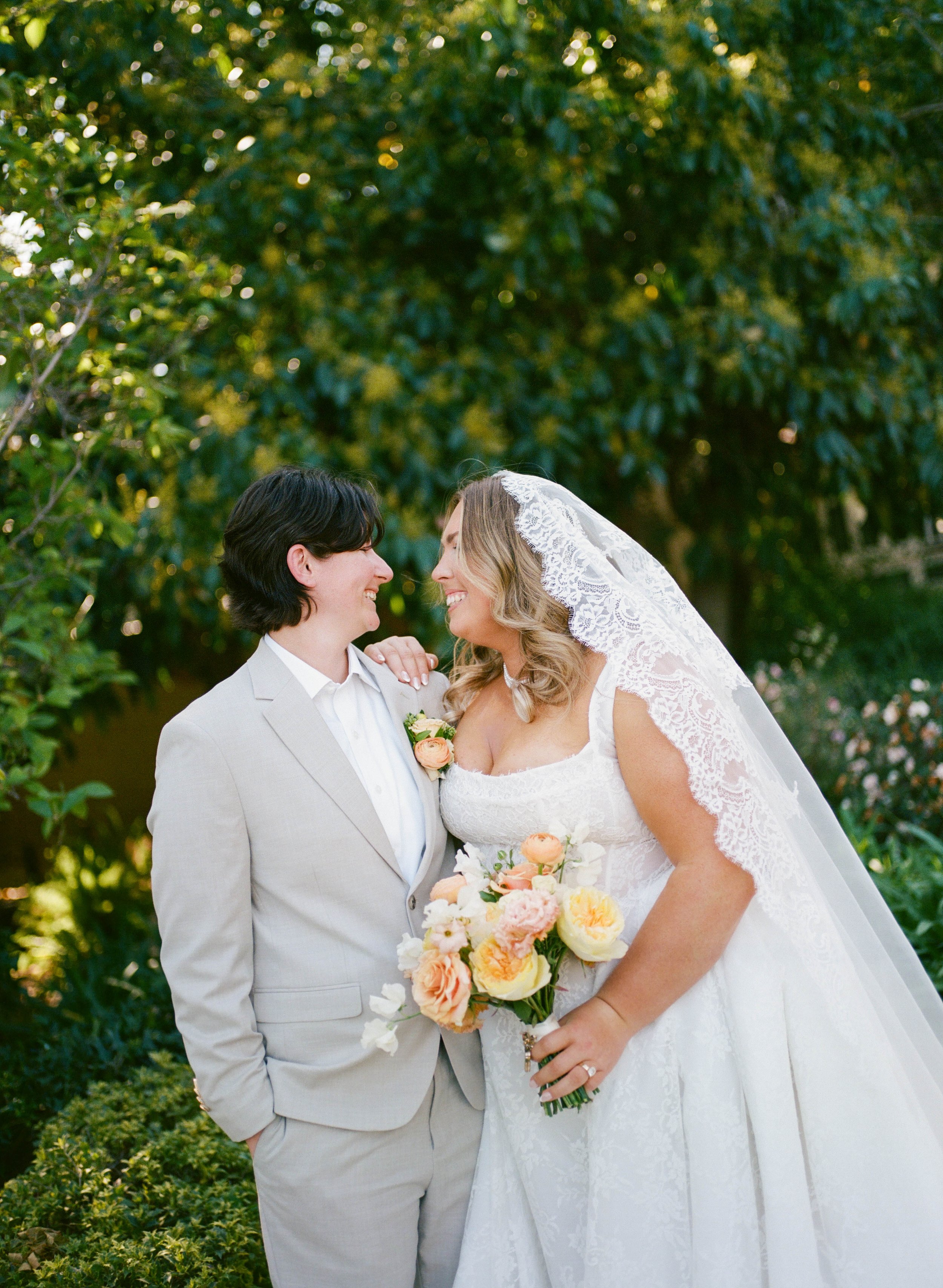A bride and groom smiling and looking at each other outdoors. The bride wears a white lace wedding dress and veil, holding a bouquet of peach, white, and yellow flowers. The groom is in a light gray suit with a boutonniere. Green foliage in the backg
