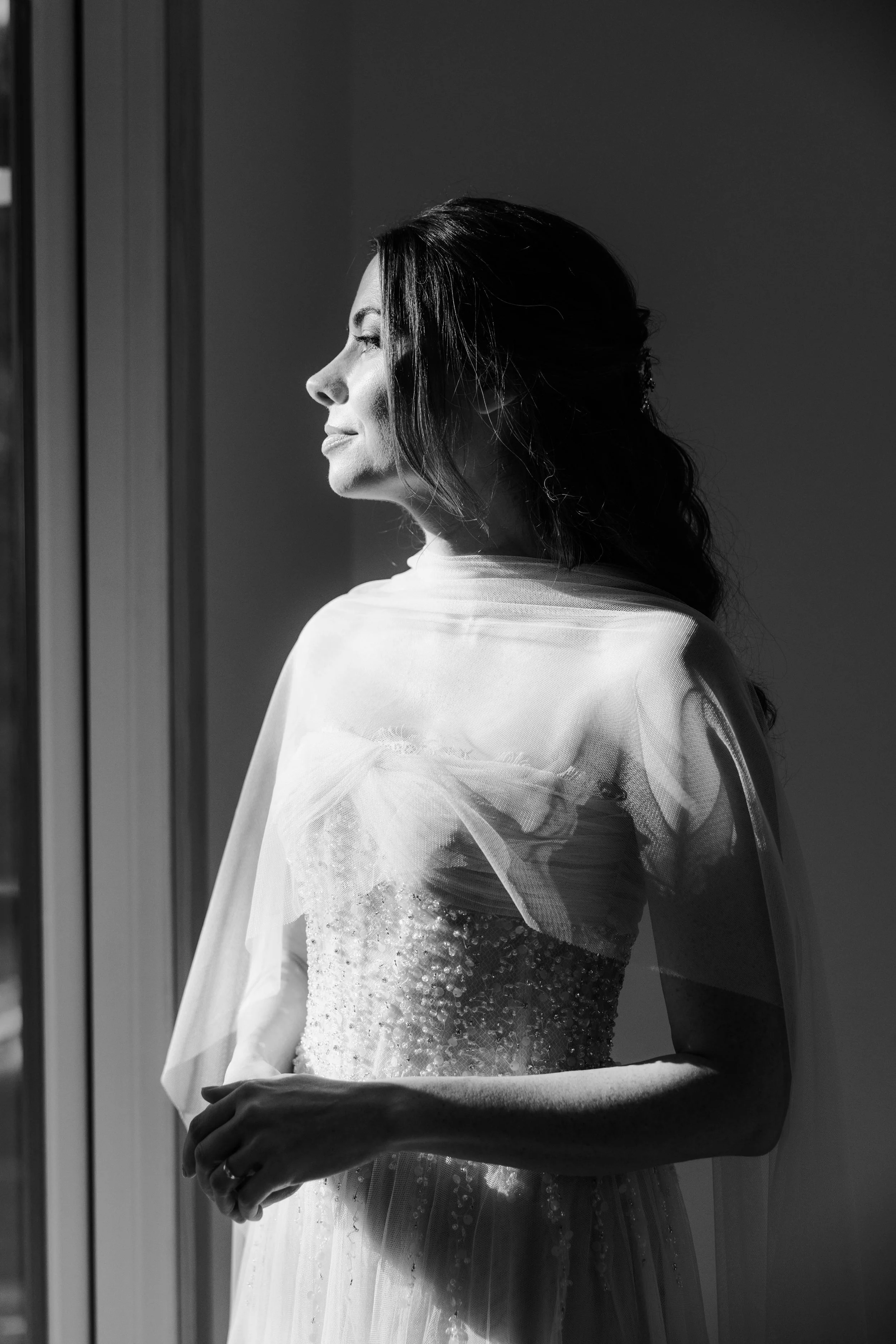 A black and white side profile of a woman in a wedding dress looking out a window.
