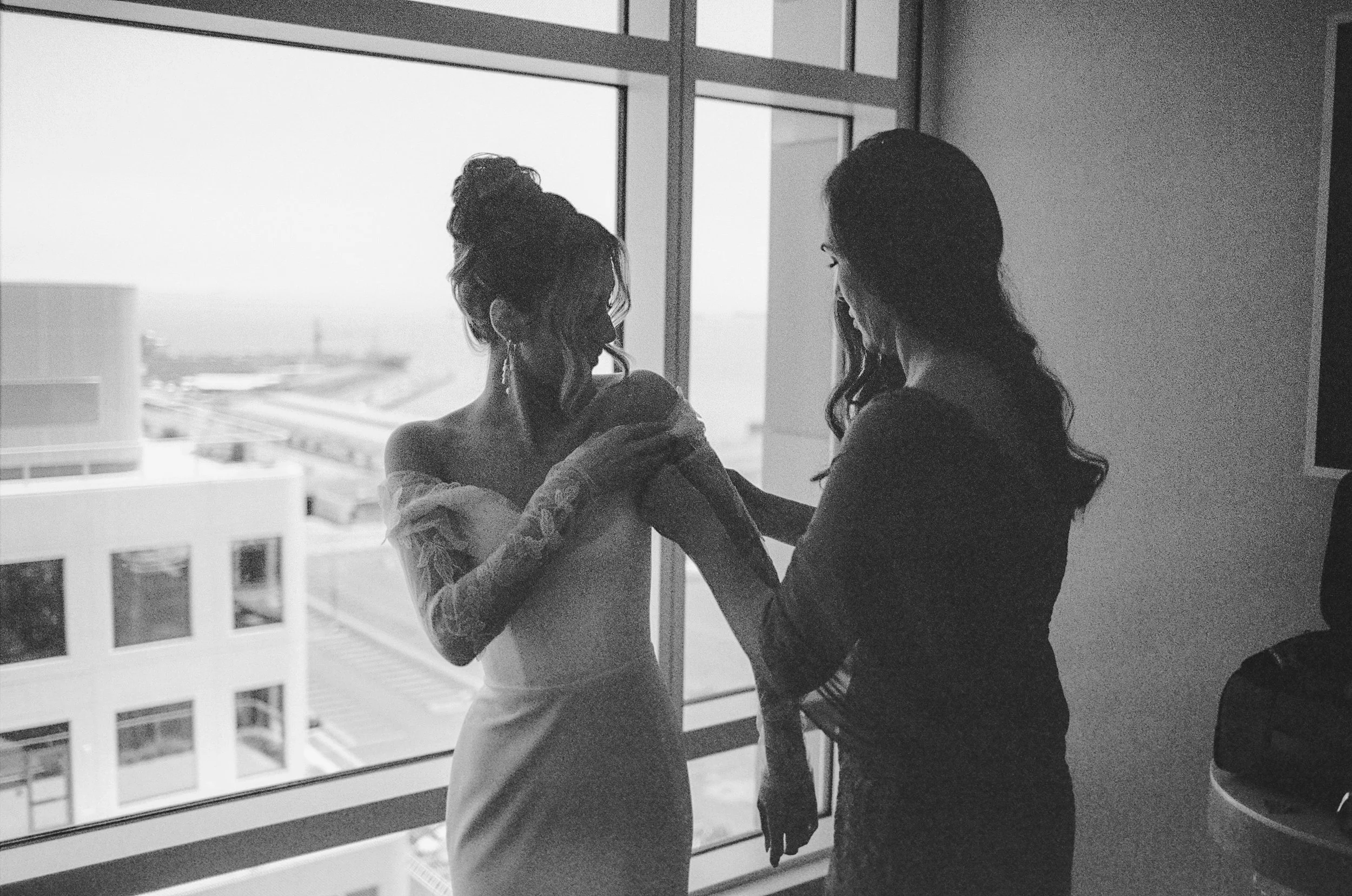 Two women with long hair interacting near a large window in a high-rise building, with cityscape view outside.