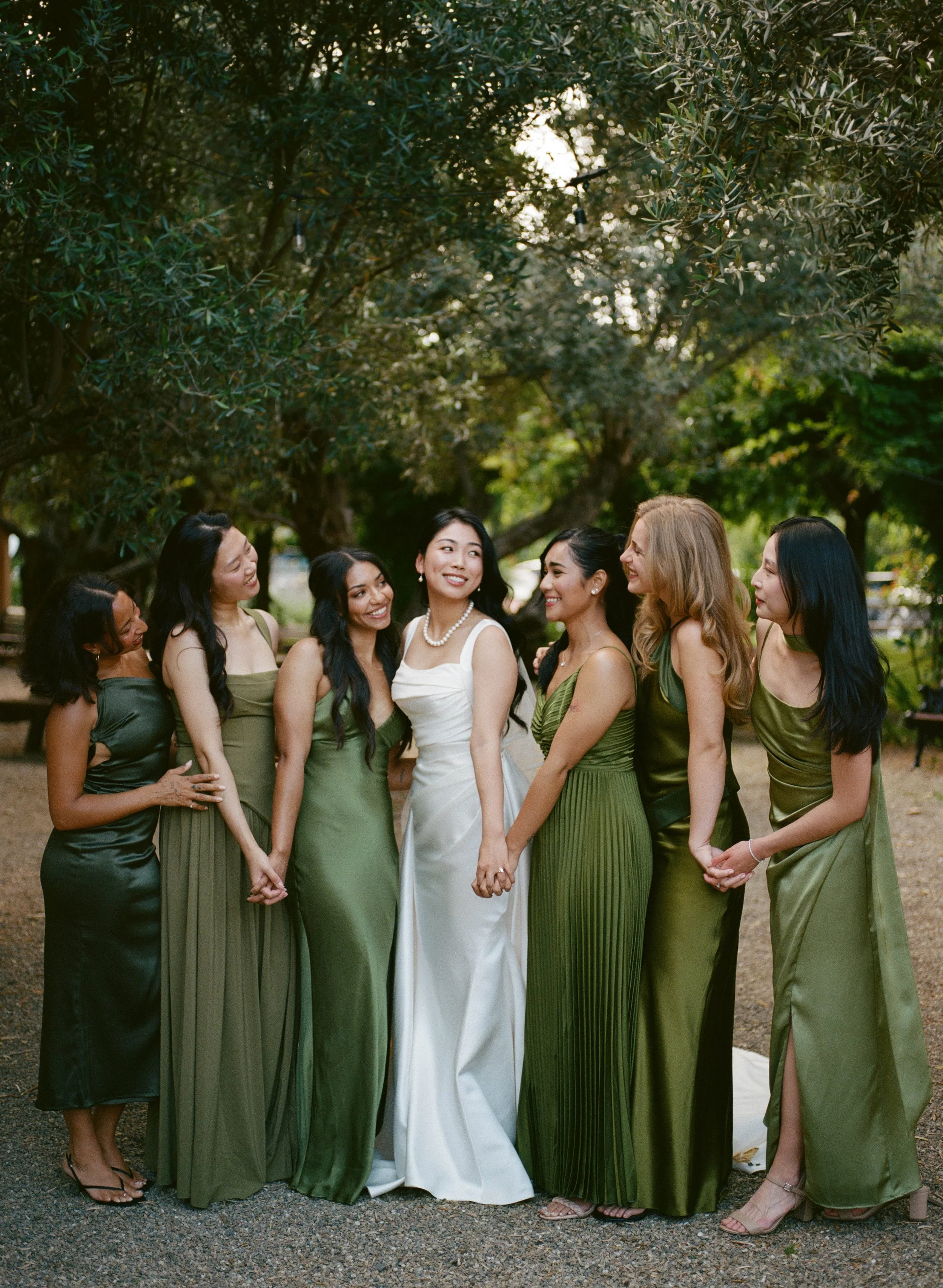A group of women dressed in elegant green dresses and one woman in a white dress, holding hands and smiling outdoors beneath trees at sunset.