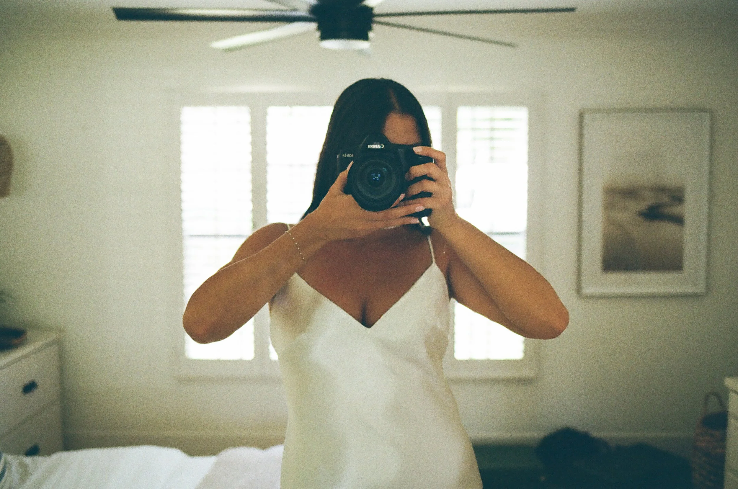 A woman taking a mirror selfie with a professional camera in a bright, minimalistic room with white walls, window shutters, and artwork on the wall.